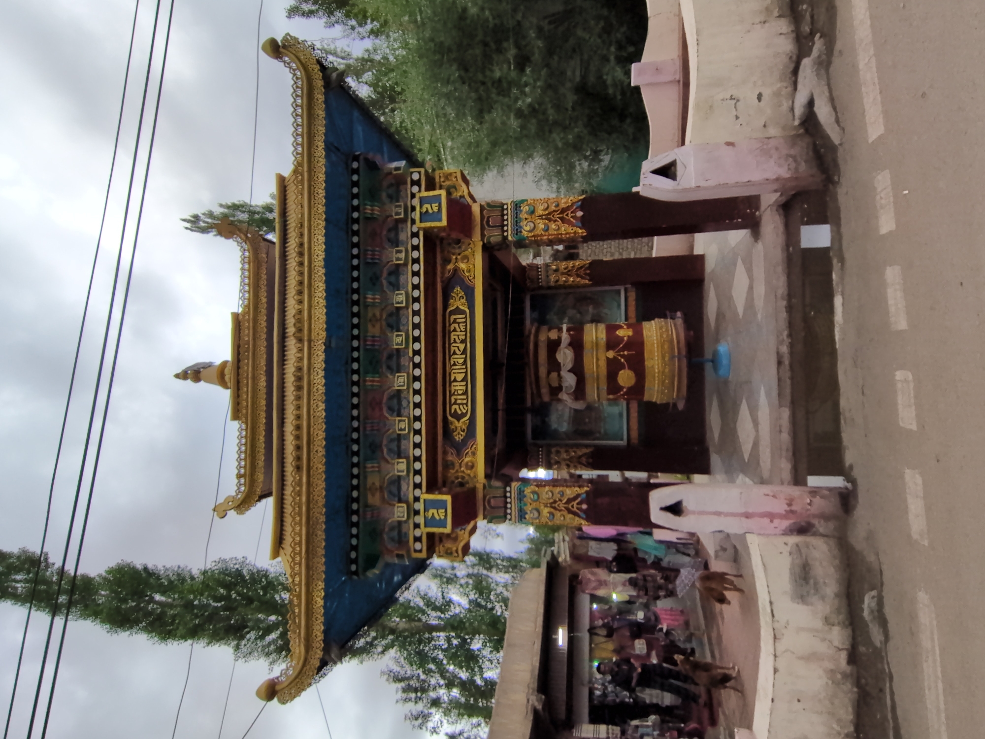 The image captures a Buddhist prayer wheel structure in the city of Leh, India. The structure features an ornate golden roof with blue detailing and intricate patterns. A large, cylindrical prayer wheel, adorned with red and gold, is centrally placed within the structure. The area around the prayer wheel is paved, and there are two dogs near a low, curved wall in the foreground. Behind the prayer wheel, a market stalls are visible with people browsing the goods. The sky is overcast, and tall trees line the background. The overall scene depicts a religious and cultural site within a bustling urban environment.