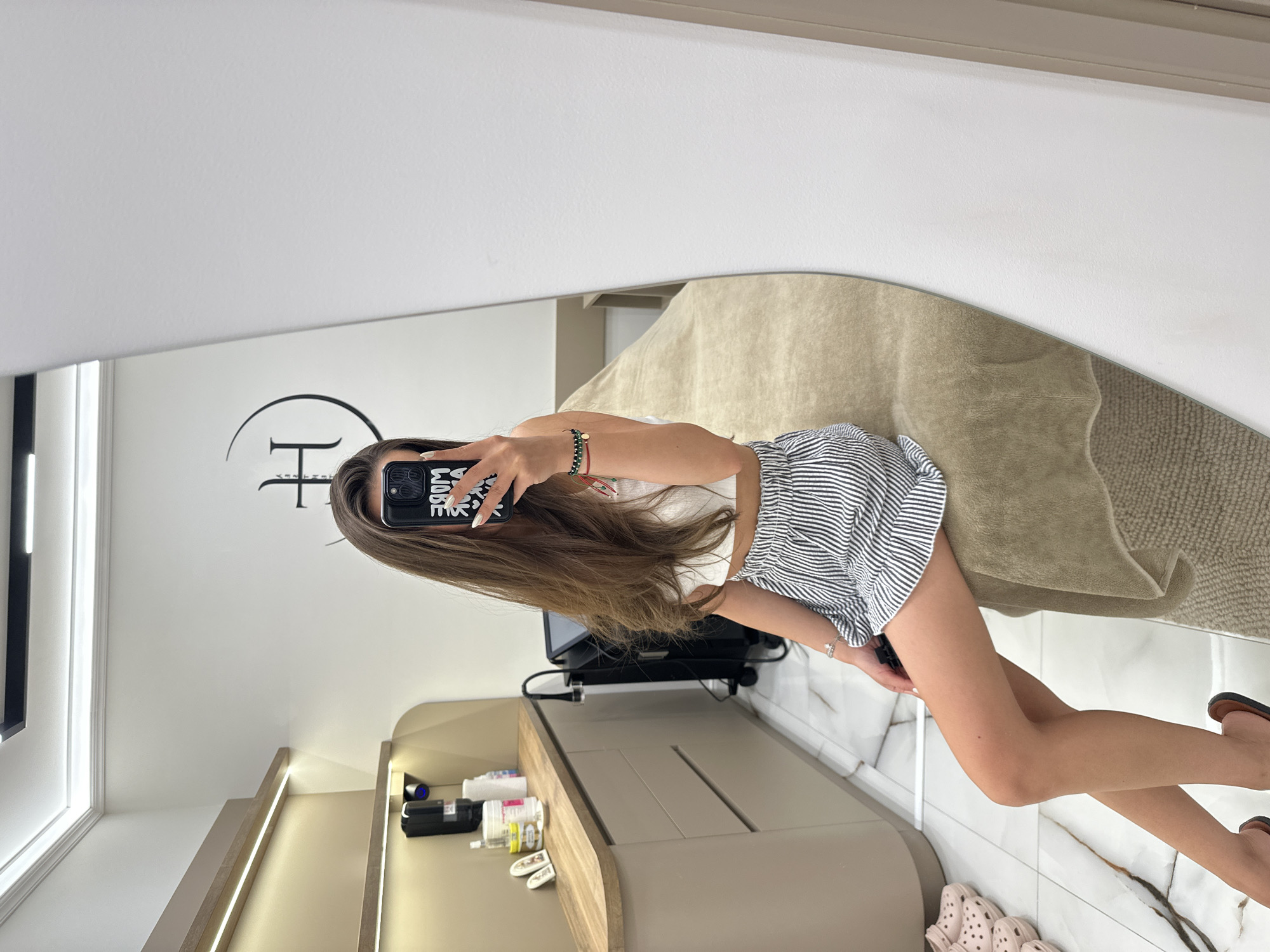 The image shows a young woman taking a mirror selfie in what appears to be a beauty salon or treatment room. She is seated on a treatment bed, wearing a white crop top and a striped skirt. Her hair is long and brown, and she is holding a phone in her right hand. The room is brightly lit, with modern, clean decor. There are shelves with products and equipment visible in the background. The floor is tiled. The logo of a beauty salon, featuring a stylized "H" inside a circle, is visible on the wall behind her. This salon is located in Petrich, Bulgaria.