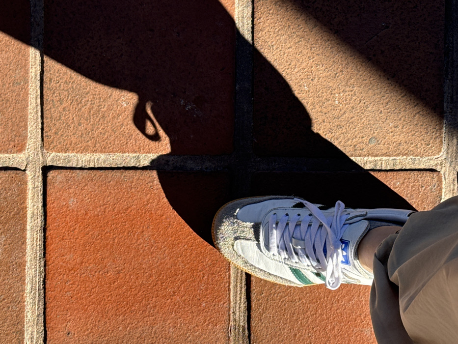 An overhead shot on terracotta paving stones. A white shoe with green stripes and white laces is partly visible, encroached upon by a large, dark shadow with an odd shape near the corner. The shadow extends over three paving stones. Beige pants are visible along the right edge, indicating the wearer's presence. Bright sunlight creates sharp contrasts, highlighting the texture of the bricks and the shoe.