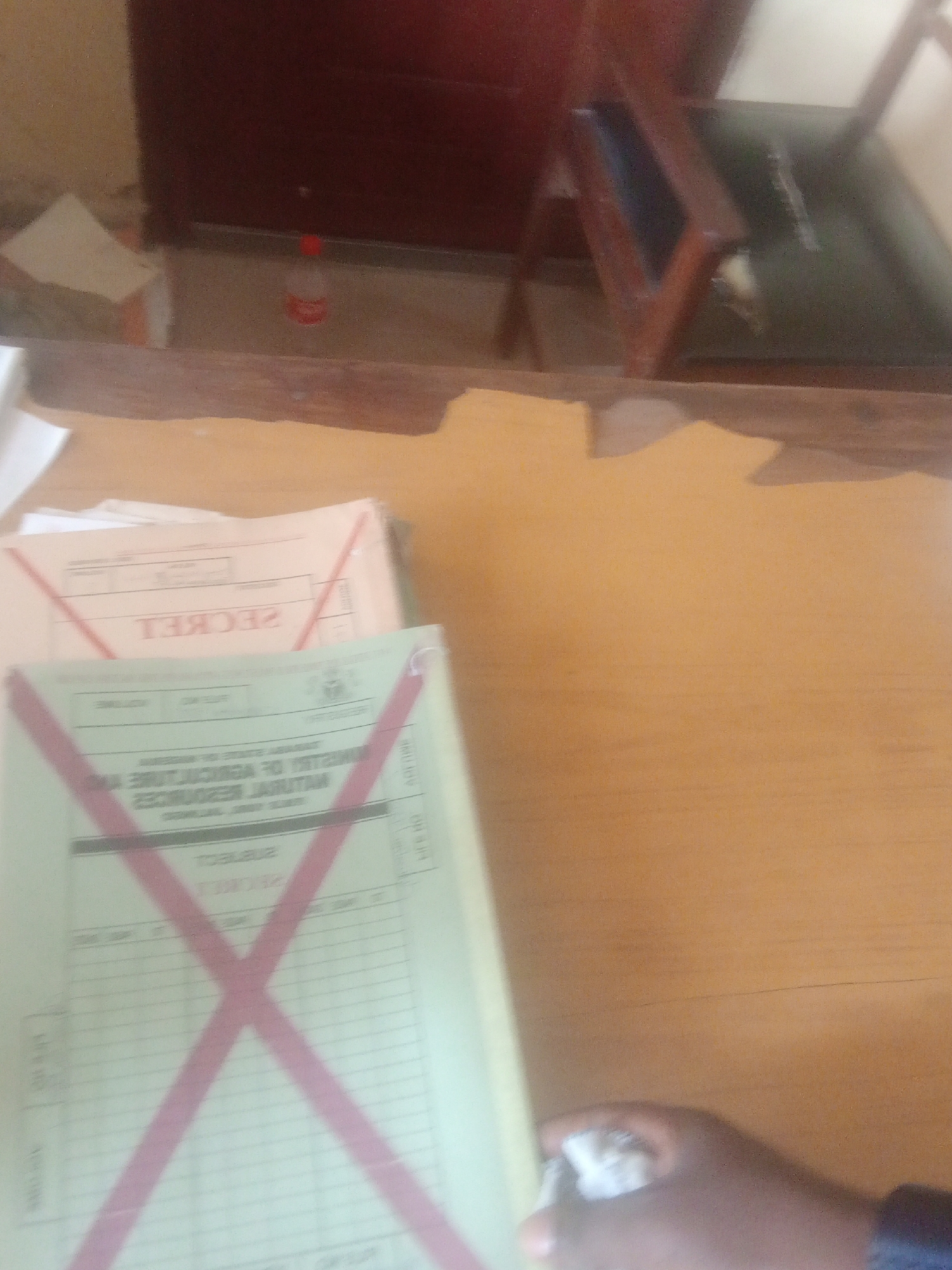 The image depicts a close-up of a wooden desk with several documents on it. Two of the documents are prominently featured, both appearing to be official forms. The top document has a red "X" marking across it and text that reads "SECRET" in reverse. The document underneath also has a red "X" marking, and some faint text is visible, suggesting it might be related to vehicle licensing or state government affairs, possibly from Nigeria given the location context. In the background, a portion of a dark red wooden cabinet or door is visible, along with a small red-capped bottle on the floor. A hand is also visible in the lower right corner, holding what appears to be a crumpled piece of paper or tissue. The scene is indoors, likely an office environment, with warm lighting that suggests daytime. The overall impression is one of administrative or official business, with some documents marked for cancellation or review.