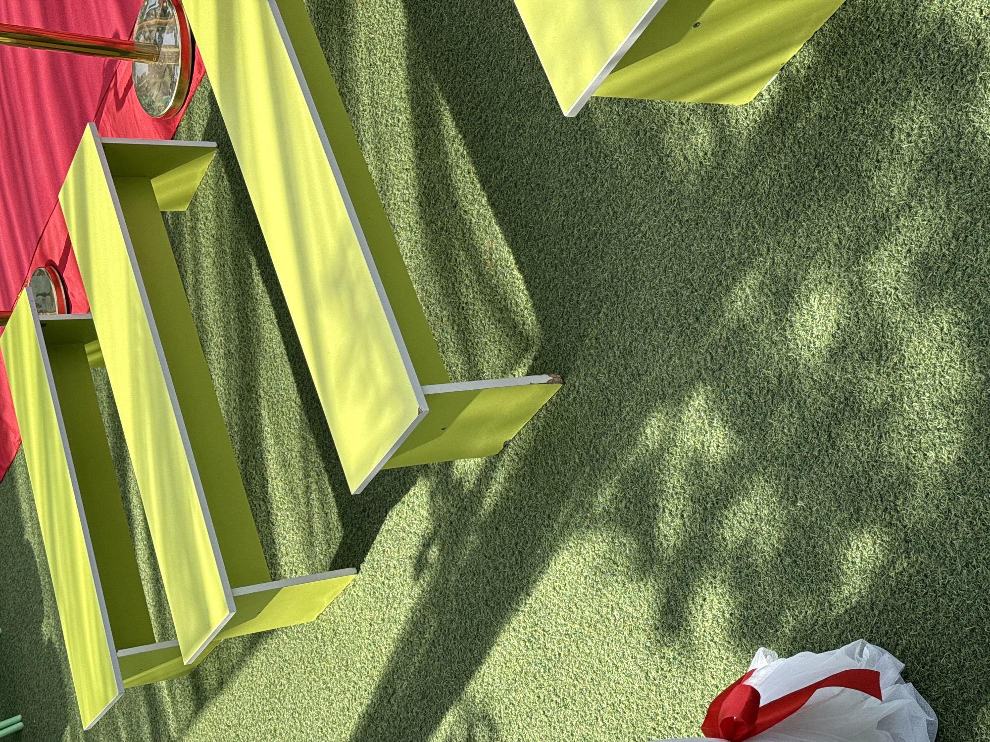 The image presents an outdoor scene featuring an artificial turf surface, overlaid with shadows of trees, and elements of a display or exhibit. Three light-green, elongated, rectangular structures are positioned on the left side of the frame, oriented diagonally. Their open-box design is accentuated by white interior edges. Adjacent to these structures, on the upper left, a portion of a red curtain and a gold-colored pole with a circular base are visible. In the lower right corner, a white plastic bag with a red ribbon accentuates the scene. The location is unidentified, making it impossible to determine the city and the state.