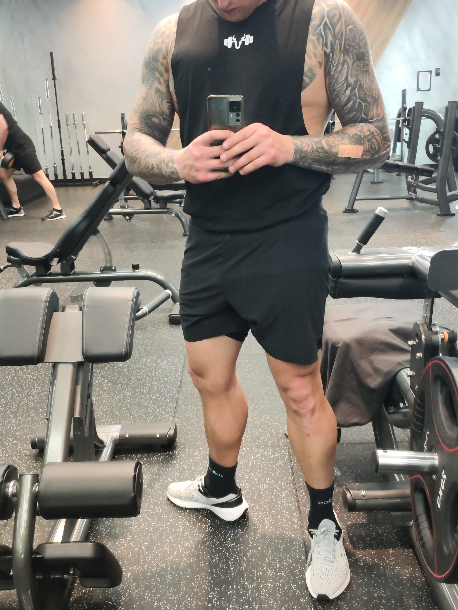 A man is standing in a gym, taking a selfie in a mirror. He is wearing a black tank top, black shorts, black socks, and grey running shoes. He has tattoos on both arms. He is holding a smartphone. In the background, there are weightlifting machines and another person exercising. The location is likely Burgas, Bulgaria, based on the user prompt, but there are no visual clues in the image that confirm this. The gym appears to be well-equipped.