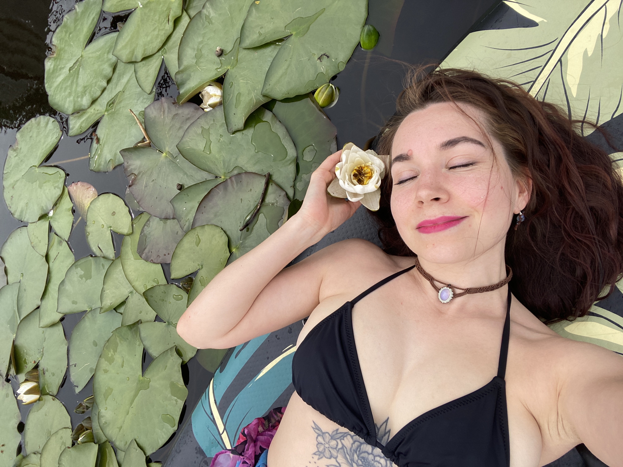 A woman with long, dark, wavy hair lies on her back on a float in a body of water surrounded by lily pads. She is wearing a black bikini top and a brown macrame choker with a light-colored stone pendant. Her eyes are closed, and she has a serene expression. Her right arm is raised, and she holds a white water lily to her temple. A tattoo of flowers is visible on her chest. The water is dark, and the lily pads are a vibrant green, some with water droplets on them. The float has a large, stylized leaf pattern on it. The lighting suggests it is daytime, likely sunny, given the brightness of the scene. The presence of lily pads and dark water indicates a natural setting, such as a pond or lake. The woman appears to be relaxing and enjoying the peaceful surroundings.