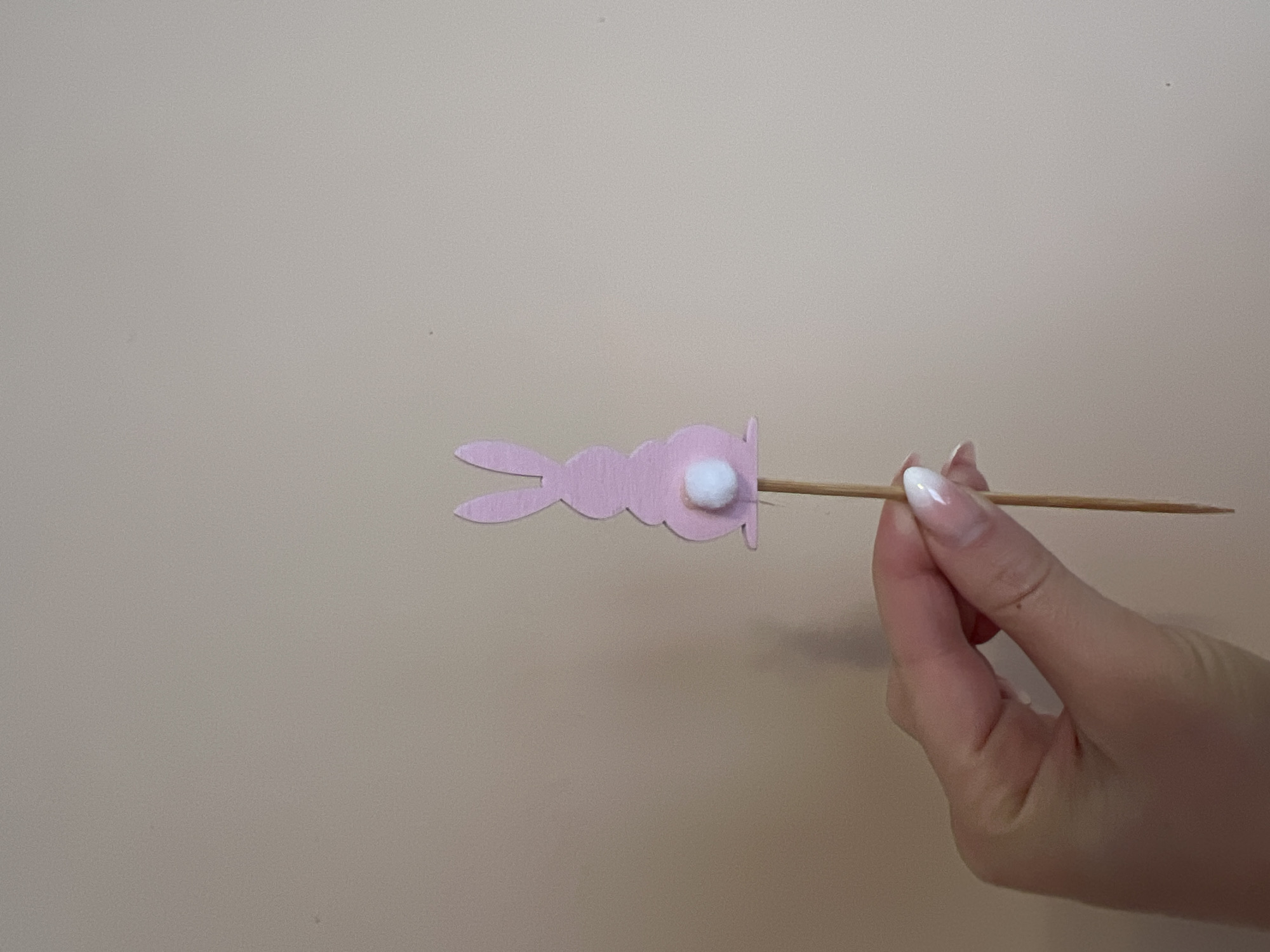 A hand holds a pink bunny-shaped craft with a white pompom tail attached to a wooden skewer against a light beige background. The scene appears indoors, with a simple, undecorated wall visible. There is no indication of the location being in Yambol, Bulgaria.