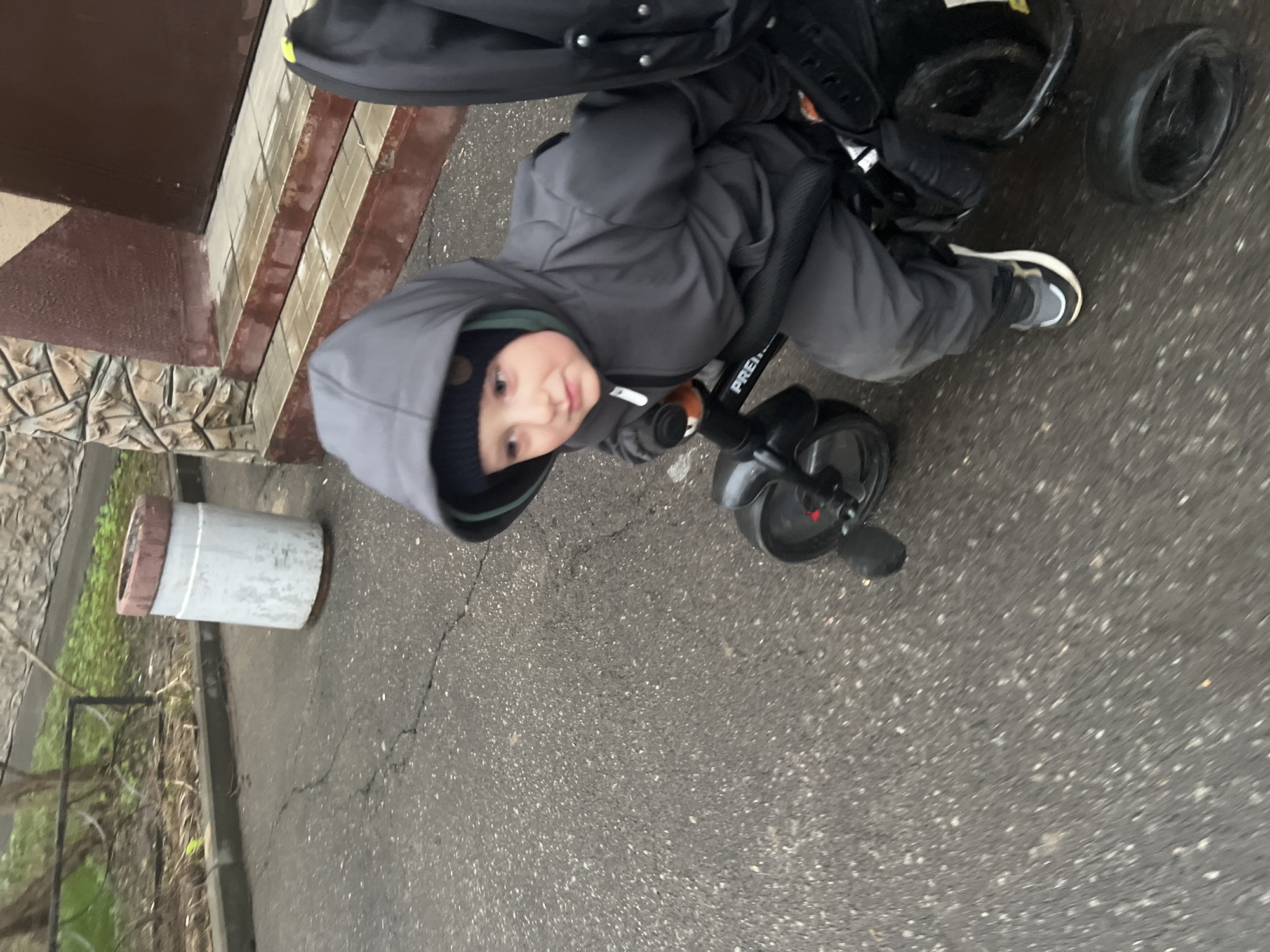 A young child in a gray hooded jacket and black beanie is seated on a tricycle on a gray asphalt surface. The tricycle is mostly black with the word "PREMIUM" visible on the handlebars. The child is looking towards the camera. To the left, a building with brown paneling and stone-like decorative elements is visible, alongside a gray cylindrical object resting on the ground. The scene appears to be outdoors, potentially on a sidewalk or paved area adjacent to a building. The weather appears overcast, and the location is suggested to be within the city of Mytishchi, Russia.