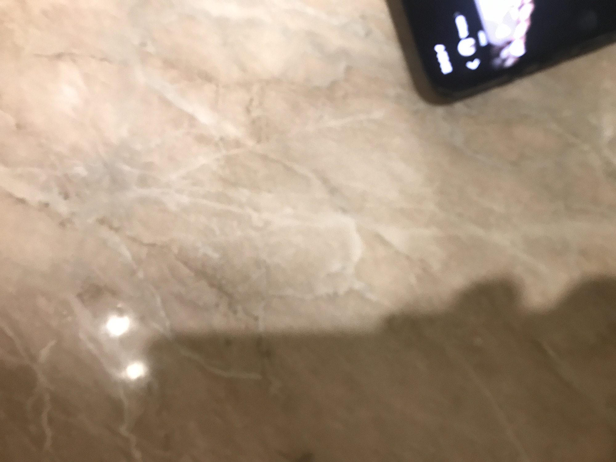 The image shows a close-up of a light brown, patterned surface, possibly a marble tabletop. A portion of a smartphone is visible in the upper right corner. The bottom right portion of the frame is dark, possibly due to a shadow. There are no indications of location or specific details about people, actions, or interactions in the scene.