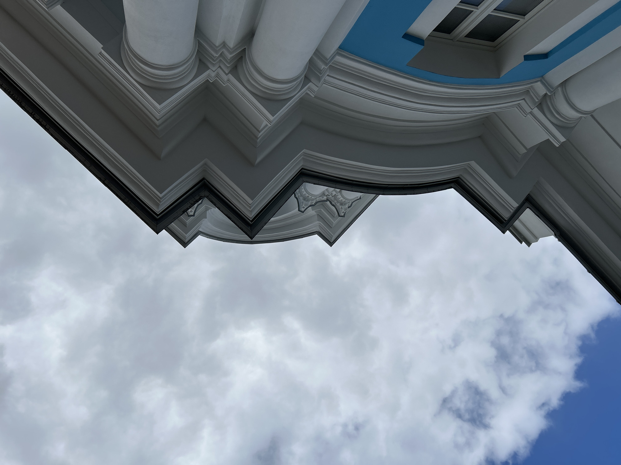 The image shows an upward view of a building exterior, featuring ornate white architectural details, including columns and layered molding. Part of a blue ceiling is visible. The rest of the frame is occupied by a cloudy sky. The building may be located in Saint Petersburg, Russia.