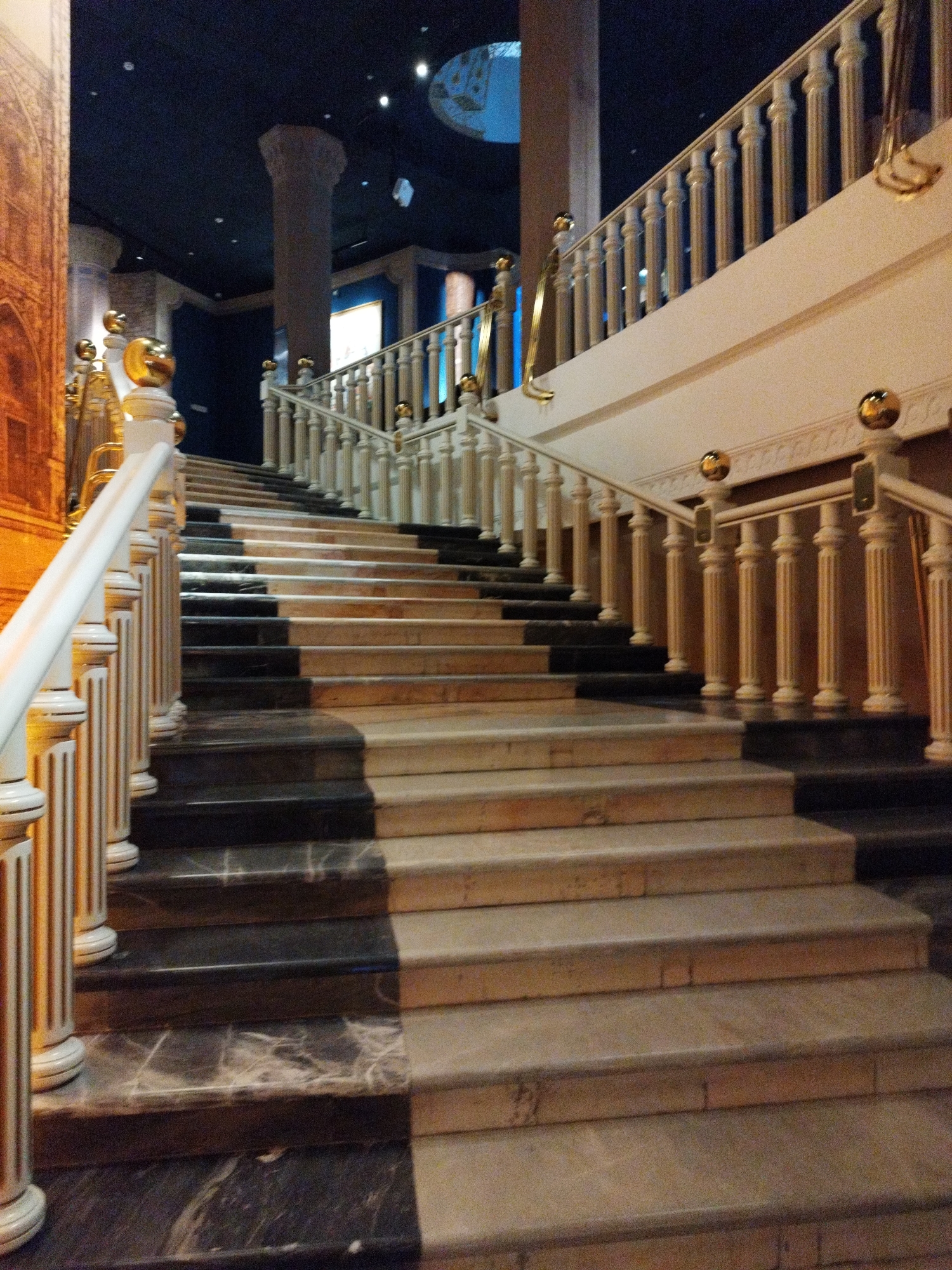 This media file captures a grand, ornate staircase, likely indoors. The staircase itself is a prominent object, featuring alternating steps of dark marble and lighter beige stone. The treads are polished, reflecting the ambient light, and the risers are the same light stone as the treads. A white balustrade with decorative gold accents lines the right side of the stairs, featuring slender, fluted balusters topped with gold spheres. A matching white handrail with gold detailing curves upwards along the balustrade.
The scene is lit with artificial light, suggesting it is either evening or an indoor environment with no natural light. The overall mood is one of elegance and opulence, likely a hotel lobby, museum, or a grand event venue. The background shows a dark ceiling with some lighting fixtures, and a large, thick column. To the far left, a portion of a wall with a patterned mural or tapestry is visible, adding to the rich and detailed aesthetic. The image focuses on the staircase, with no people or active events depicted, presenting a static yet impressive architectural view. The time of day is not discernible, but the lighting suggests it is not daytime. The location is unknown, but the design implies a place with a significant architectural or decorative emphasis.