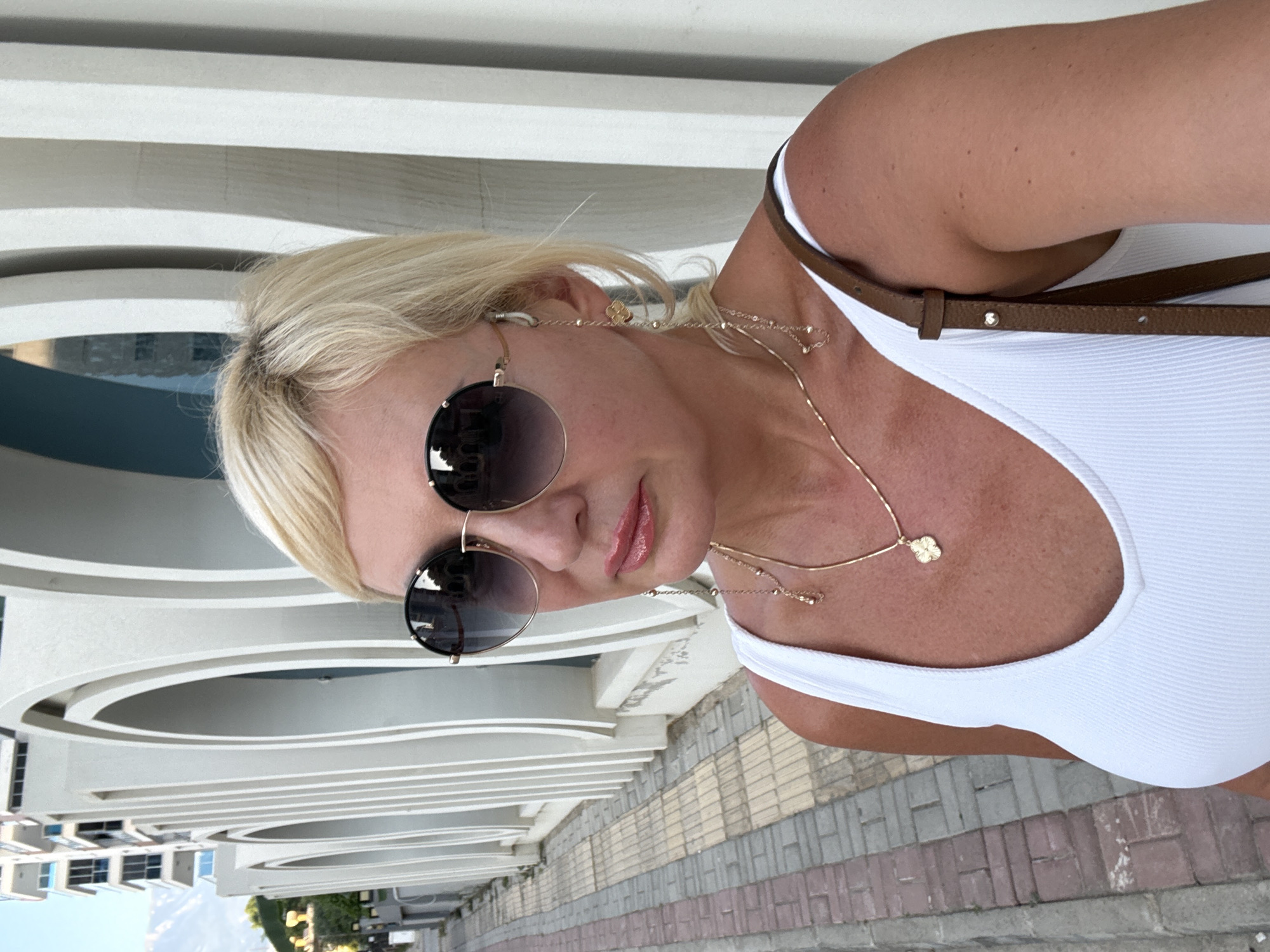 A woman with blonde hair and sunglasses takes a selfie outdoors. She is wearing a white tank top and a brown strap across her shoulder. A gold necklace with a clover pendant is visible. The background consists of a modern building with curved white architectural elements and a paved walkway. The lighting suggests it is daytime, possibly late afternoon, with bright sunlight. The overall mood appears relaxed and casual, consistent with a vacation setting. The location context is Alanya, Turkey.