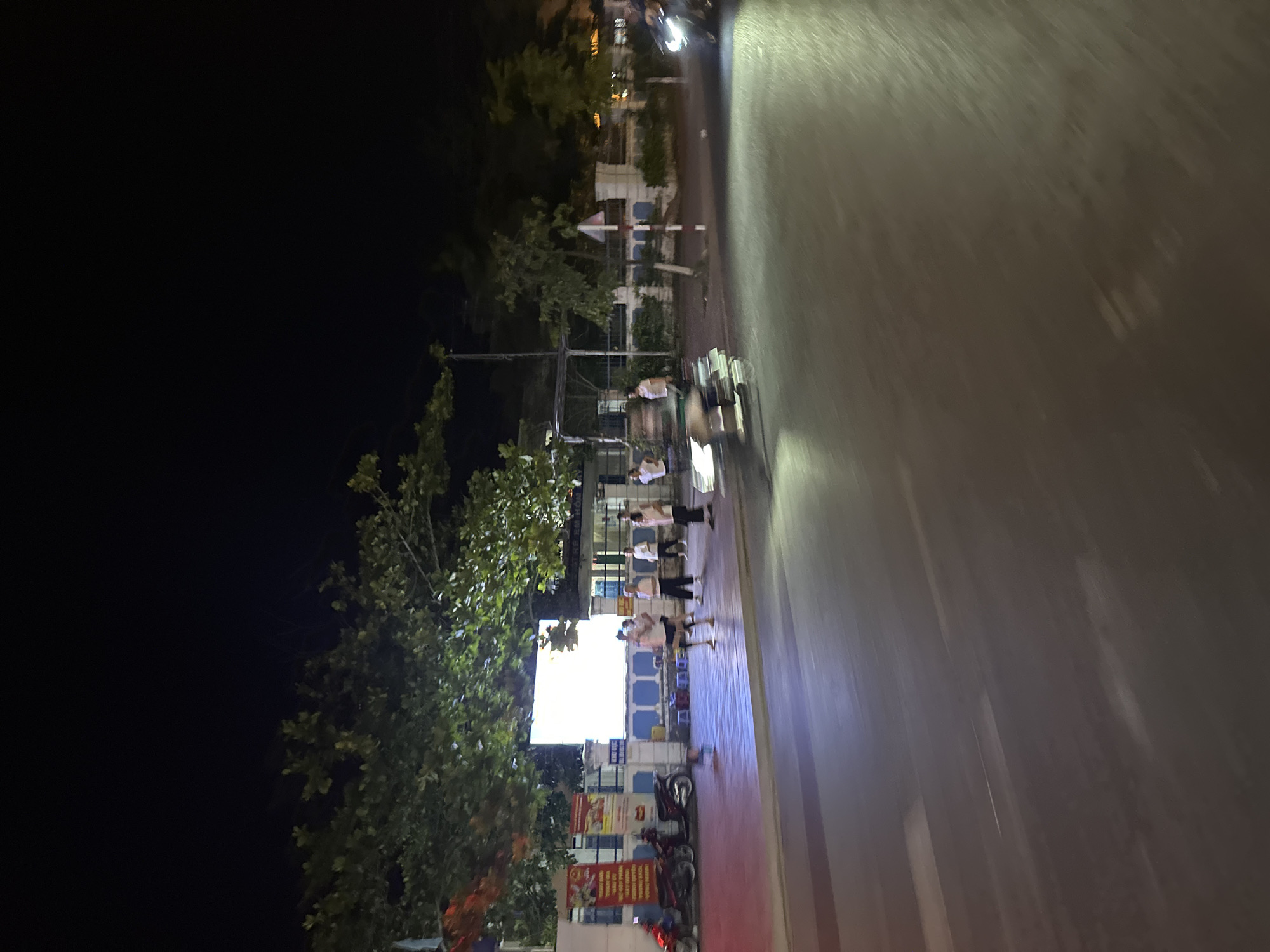 The image depicts a nighttime scene in Bac Nha Trang Ward, Vietnam. A group of people stand on a sidewalk near a brightly lit storefront, with motorcycles parked nearby. Trees line the sidewalk. A road occupies the right side of the frame, with vehicles blurred due to motion. The sky is dark.