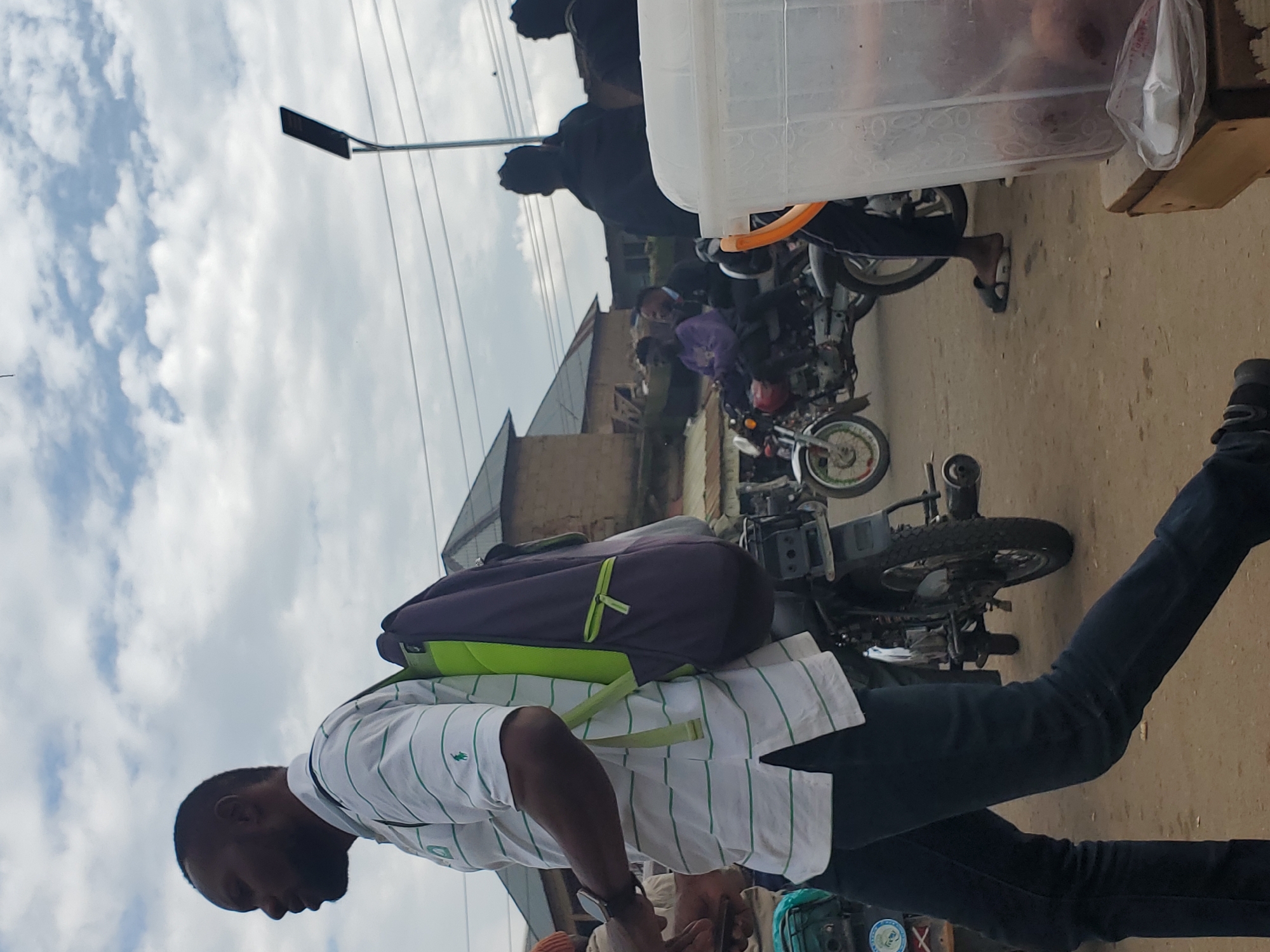 A man wearing a white and green striped polo shirt and dark pants walks from left to right, carrying a purple and green backpack. He is on a dirt road with several motorcycles parked or in motion in the background. To the right, a translucent plastic container sits on a wooden stand, holding dark-colored items. Buildings with corrugated metal roofs are visible in the distance. Power lines and a modern street lamp are also present in the background, suggesting an urban or semi-urban environment, possibly Gwong, Nigeria. The sky is partly cloudy.