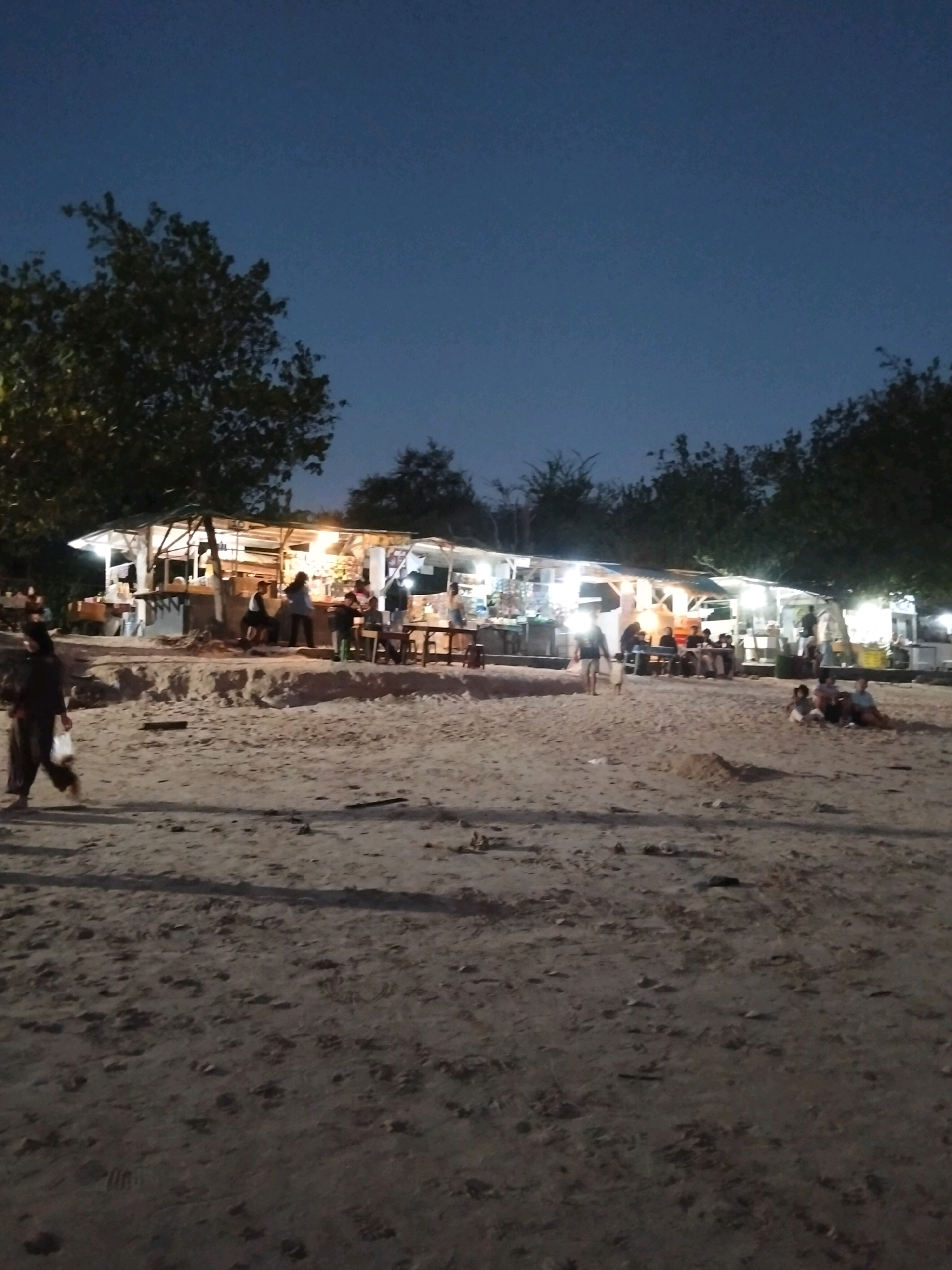 The image depicts a lively scene at a beachside food stall area in Kuta, Indonesia, during twilight. The sky is a deep blue, suggesting the sun has set. In the background, several open-air food stalls are illuminated with bright lights, showcasing goods and creating a warm glow. People are gathered at the stalls, some standing and others seated at tables, indicating a bustling atmosphere. A woman in dark clothing walks across the sandy beach in the foreground, carrying a plastic bag. The beach itself is covered in sand with some debris and footprints visible. Trees with dense foliage are scattered behind the stalls, framing the scene. The overall impression is one of a popular evening spot where locals and possibly tourists are enjoying food and the beachside ambiance.