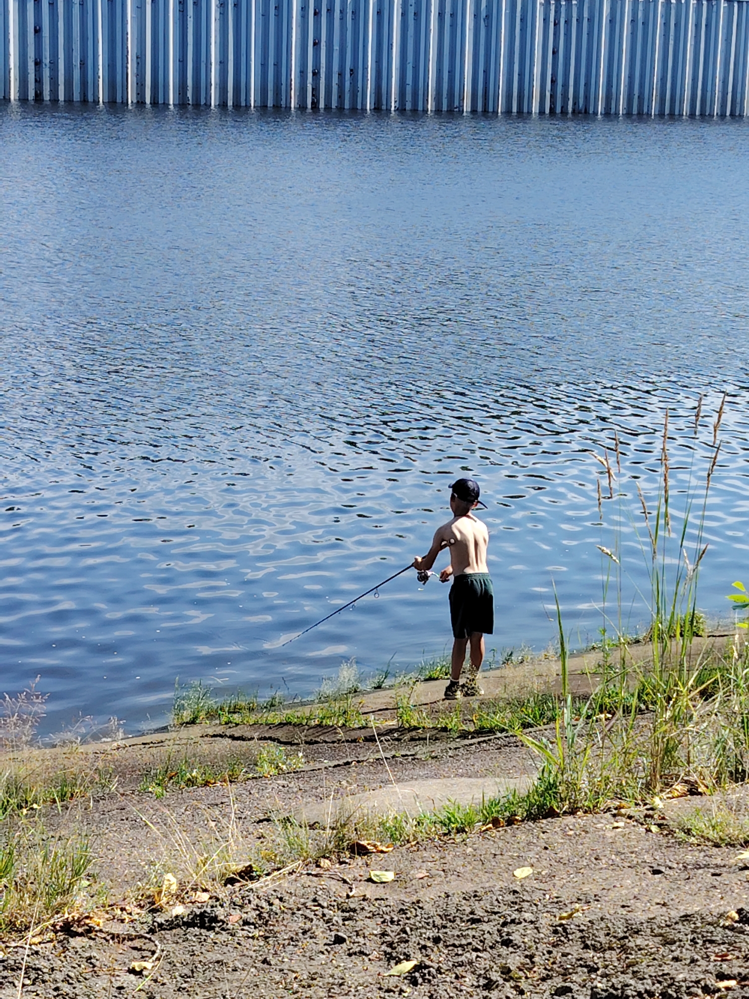 An undated, eye-level, full-shot photograph captures a young boy fishing from the bank of a body of water. The boy, with his back to the camera, stands on a gravelly shore, wearing a dark baseball cap and dark shorts. His arms are raised as he holds a fishing rod, its line extending into the rippling blue water. Behind the water, a corrugated metal barrier runs horizontally across the frame, suggesting an industrial or structural edge to the waterway, possibly a canal or reservoir. The sunlight casts a bright, clear light on the scene, highlighting the texture of the gravel and the shimmer of the water. The presence of a fishing rod and line indicates the action of angling, while the boy's stance suggests a focused or patient demeanor. The background offers no identifiable landmarks, but the image appears to be taken during daylight hours, likely in a setting with sparse vegetation along the water's edge.