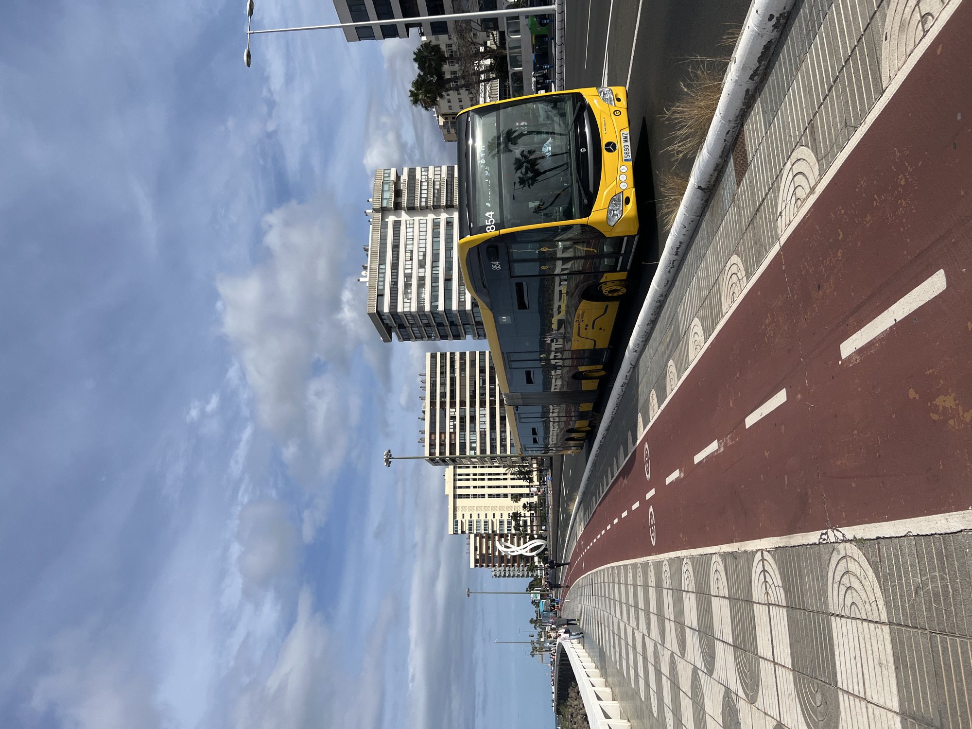 The image captures a bright yellow bus on a city street in Las Palmas, Spain. The bus is positioned to the right of a red bike lane, separated by a low concrete barrier with a semicircular design. The license plate is visible, but the details are difficult to discern.
Behind the bus, modern multi-story buildings rise into a sky dotted with clouds. Street lights line the street, and in the distance, people are visible along the pathway. The overall scene suggests a coastal urban environment on a clear, sunny day.