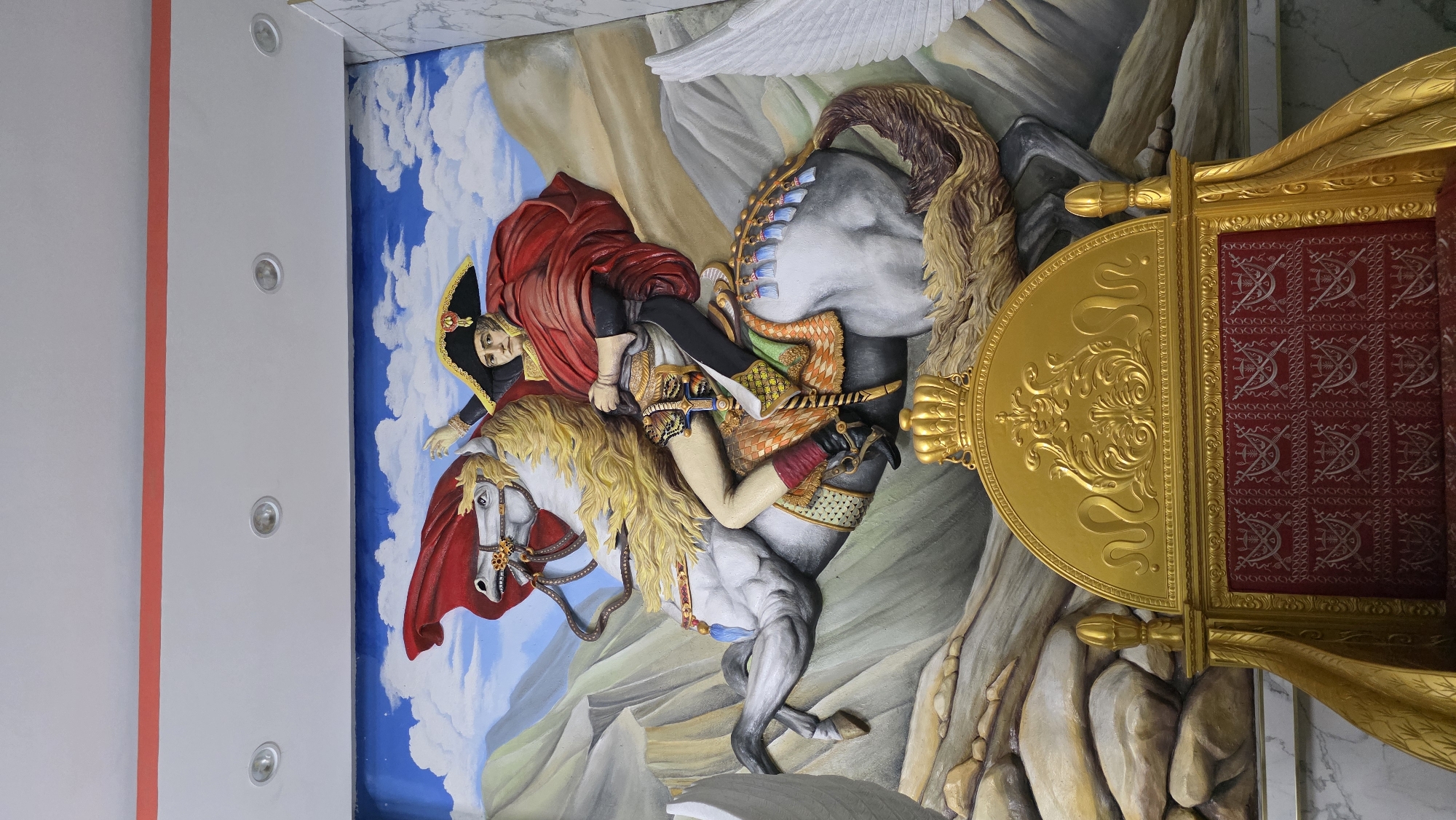 This media file displays a three-dimensional relief sculpture, likely installed on a ceiling or high wall, depicting a historical scene. The central subject is a man resembling Napoleon Bonaparte, clad in a red cape and a black bicorne hat, riding a rearing white horse with a flowing blonde mane and tail. The man's right arm is outstretched, pointing forward. The horse's body is mostly white and grey, with ornate golden and red saddle details. Another horse or part of the same one is visible, showing a grey body and a brown, bushy tail.
The background of the sculpture features a blue sky dotted with white clouds on the left and stylized rocky mountains or terrain on the right. A prominent golden, ornate structure resembling a throne's backrest or a bed's headboard is visible on the far right, upholstered in red fabric with a repeating pattern of crossed arrows or spears and bows.
The overall setting appears to be an opulent indoor space. Adjacent to the sculpture on the left is a plain white ceiling or wall section with three recessed silver-rimmed lights and a thin vertical red stripe. The artwork's style is grand and theatrical, conveying a sense of power and heroism through the dynamic pose of the rider and horse. No visible text is present in the image.
