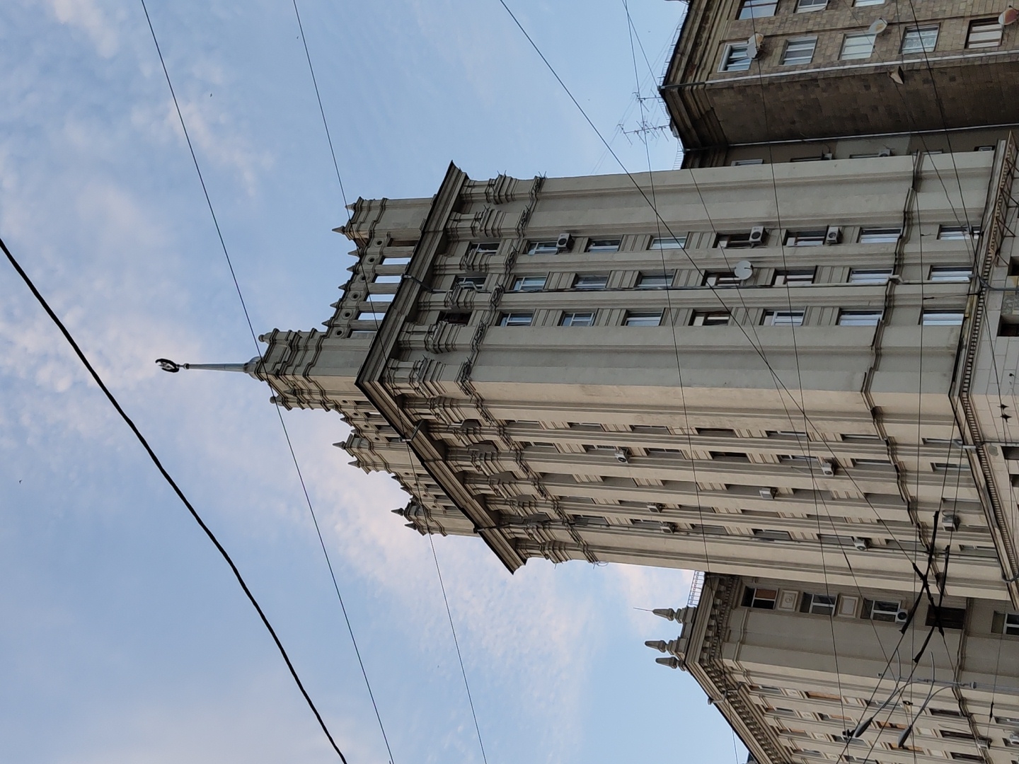 A low-angle shot captures the upper floors of the Ministry of Infrastructure building in Kharkiv, Ukraine. The pale beige building, constructed in the Stalinist Empire style, dominates the frame with its symmetrical design, pilasters, and ornate cornices. A prominent spire topped with a red star and crescent moon is visible at the apex of the central tower. Multiple wires, likely for trolleybuses or trams, crisscross the sky and obscure parts of the building. The sky is a pale blue with wispy clouds. To the right, the darker, grittier facade of an adjacent apartment building peeks into view, contrasting with the grander architectural style of the ministry. No people are visible in the image.