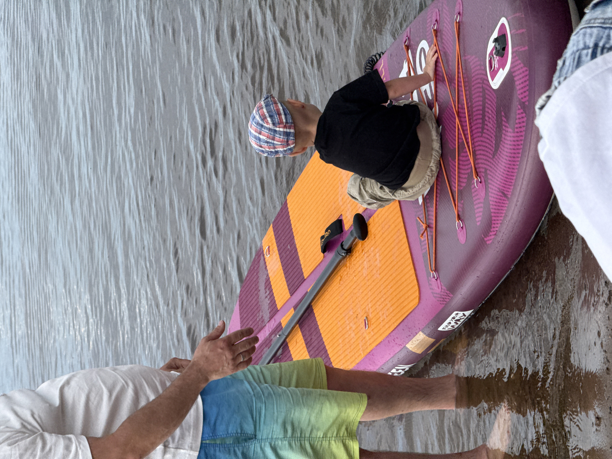 The image depicts a scene at a body of water, likely a river or lake, in Chaikovsky, Russia. In the foreground, a person is standing in shallow water, wearing a white t-shirt and gradient shorts. Their left hand is extended towards a purple paddleboard with orange accents, which is partially submerged. On the paddleboard, a young child is seated facing away from the camera, wearing a black t-shirt and tan shorts, and a red, white, and blue plaid cap. The paddleboard has orange bungee cords stretched across its surface. The water is calm with visible ripples. The overall scene suggests a recreational activity taking place outdoors.