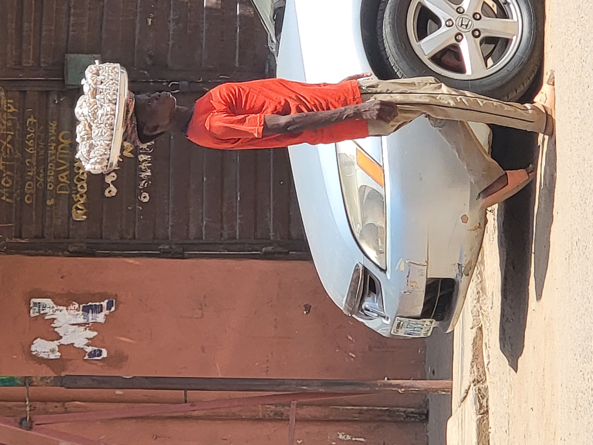 A man carrying a large bundle of goods on his head walks past a silver car in what appears to be Maiburiji, Nigeria. He wears a red long-sleeved shirt, tan pants, and sandals. The background features a weathered orange wall with peeling posters and graffiti. A closed metal gate is also visible. The front of the car is slightly damaged. The scene is well-lit with direct sunlight.