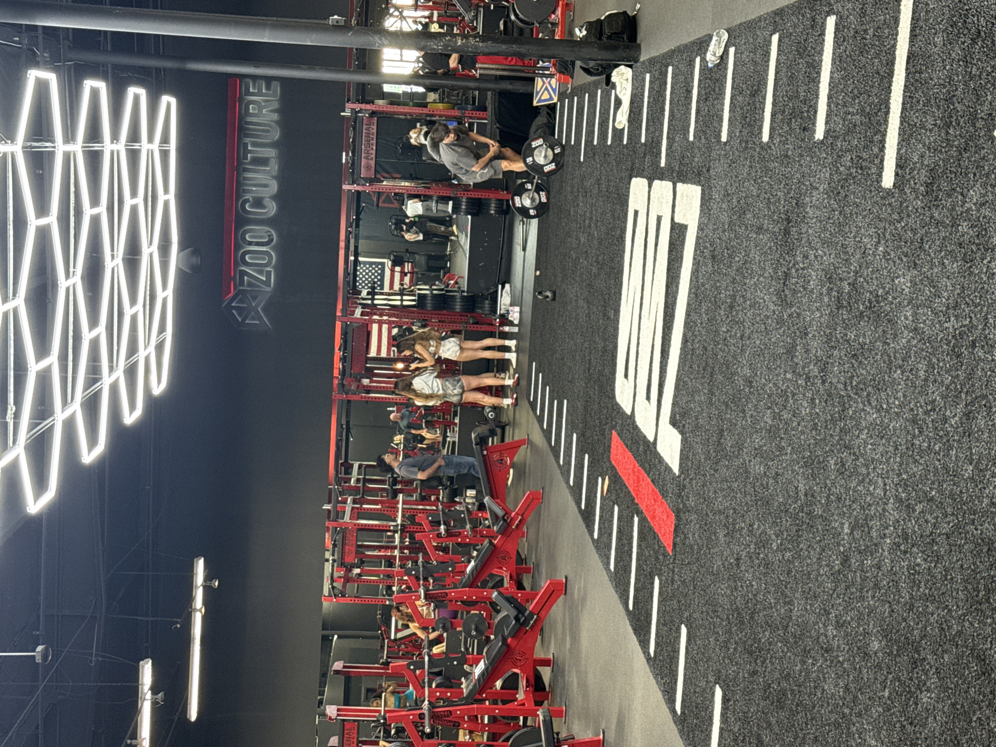 This image captures the interior of a modern gym called "ZOO CULTURE," located in Los Angeles, USA. The setting features a dark aesthetic with black walls and a dark gray carpeted floor marked with white lines, reminiscent of a sports field, including a prominent white "00" (or "100") and a red line.
Several people are present, engaged in strength training. In the foreground, a man in a gray shirt and shorts is seen lifting a barbell with heavy weights, specifically plates marked "200," suggesting deadlifts or squats. Further into the gym, two women in athletic wear stand near a row of equipment. Other individuals are visible in the background, interacting with the red strength training racks and benches, many of which bear the "ARSENAL STRENGTH" brand name.
The gym's decor includes a large, illuminated "ZOO CULTURE" sign on a black wall, featuring a stylized claw mark through the "ZOO" text. Geometric, honeycomb-patterned LED lights brightly illuminate the space from the ceiling, alongside other linear light fixtures. An American flag is visible hanging on one of the red equipment racks. A plastic water bottle rests on the floor near the white line markings. The overall atmosphere is focused on intense physical training within a well-equipped, purpose-built facility.