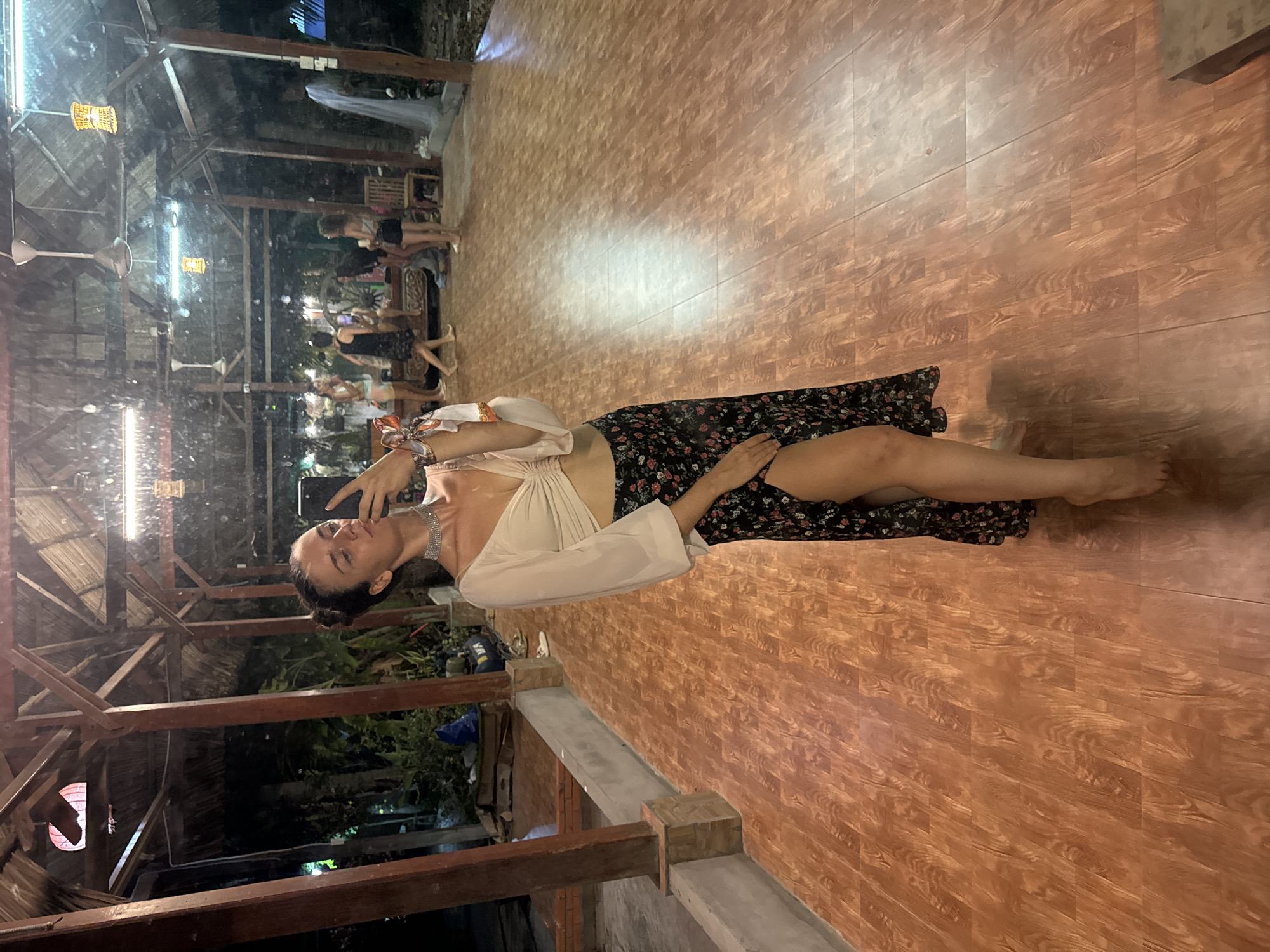 A woman is posing for a selfie in Phu Thuy Ward, Vietnam. She is wearing a white top and a black floral skirt. The background includes a wooden floor, a building with exposed wooden beams, and other people in the distance.