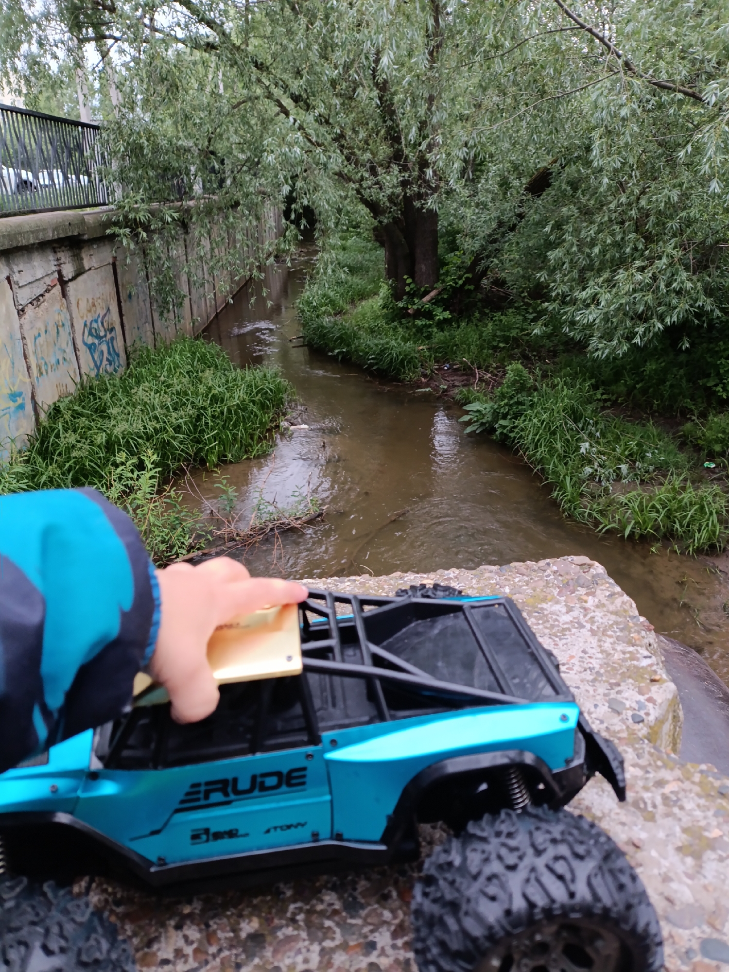 A person's hand touches the gold-colored roof of a blue "Erude" toy car parked on a concrete ledge overlooking a small stream. The stream flows between concrete walls on one side with graffiti, and grass, bushes, and trees on the other side. A metal railing is visible along the wall. The photo was taken in Chaikovsky, Russia.