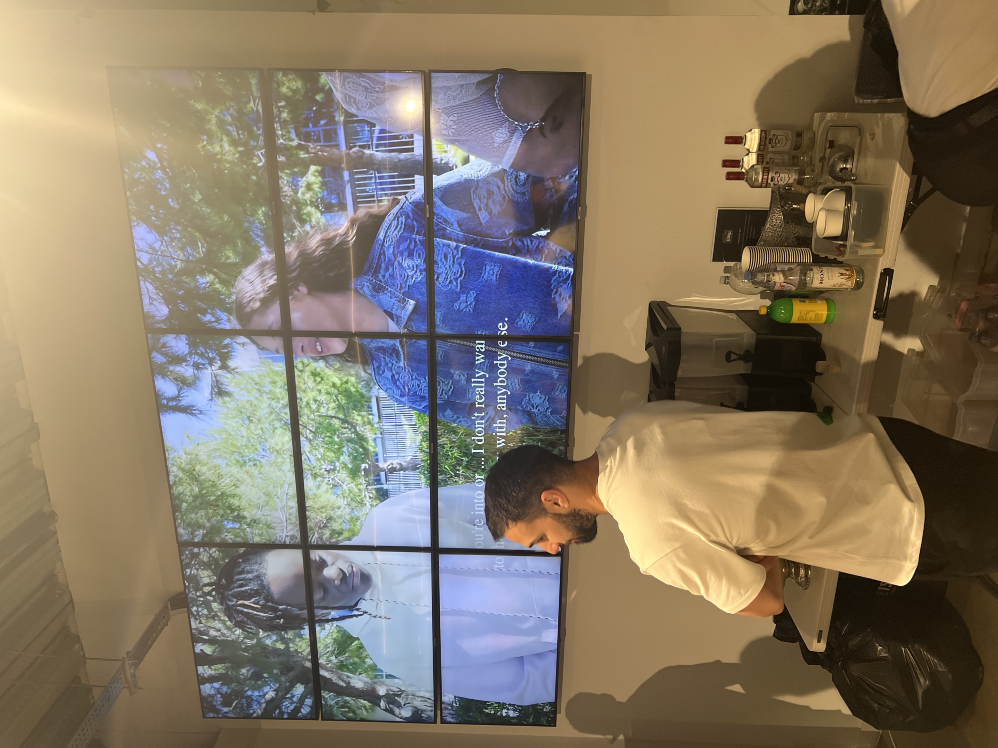 The image captures an indoor setting, likely a casual event or a modern workspace, featuring a large video wall, a person, and a beverage station. The image is rotated 90 degrees counter-clockwise.
The focal point is a large video wall composed of multiple screens displaying a mosaic of images and video clips. The content on the screens shows various individuals, including a person with dreadlocks and another with long, wavy hair, possibly a woman, in a denim jacket. Some frames show lush green foliage, suggesting an outdoor or natural theme. Overlaying some of the video segments is text, partially visible, which reads: "ou're into or ... I don't really wa" and "with, anybody e se." This likely forms a sentence such as "you're into or... I don't really want... with, anybody else."
In the foreground, a person with a beard, wearing a white t-shirt and dark pants, is standing. Their back is mostly towards the viewer as they lean over, seemingly engaged with a laptop or similar device placed on a surface. A black trash bag is visible near their feet.
To the right of the person and the video wall, a makeshift beverage station is set up on a white table. It holds several items including multiple bottles of Smirnoff vodka, a bottle of Monin syrup, a bottle of what appears to be lemon juice, and various plastic and paper cups. A small silver ice bucket or container is also present, along with a black booklet labeled "Εδέμ".
The lighting in the room appears to be artificial, with a bright lens flare emanating from the upper left portion of the video wall. The overall atmosphere suggests a relaxed, possibly social, environment where visuals and refreshments are provided.
