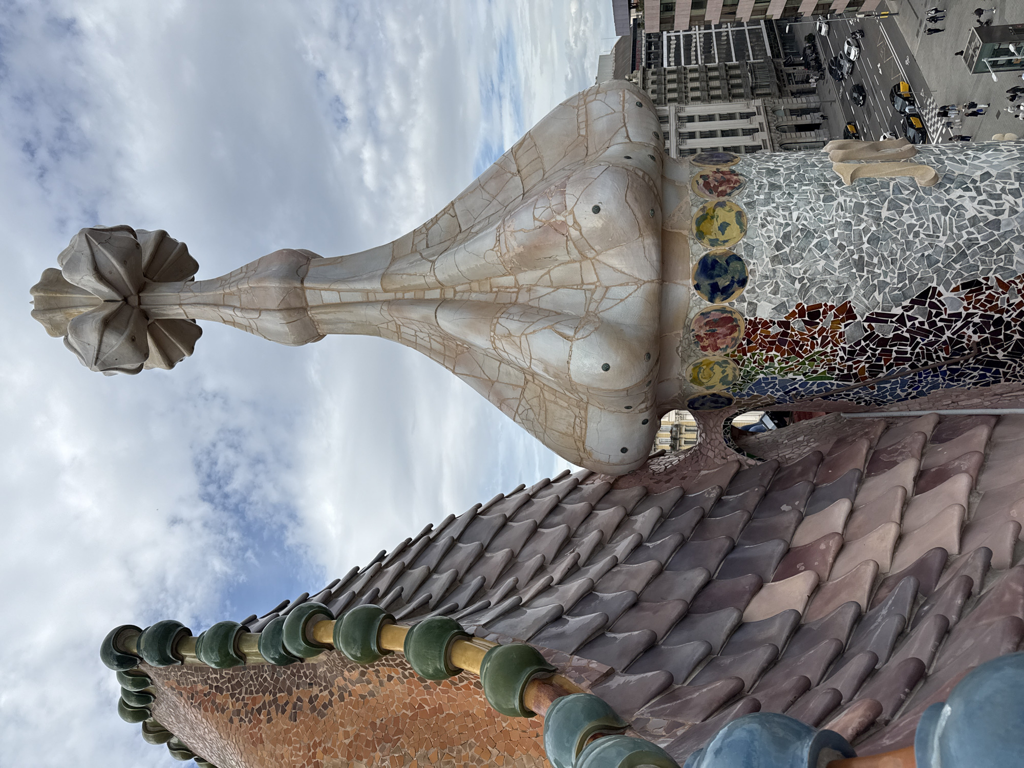 The image displays an elevated view of distinct architectural elements from the rooftop of Casa Batlló, situated on Passeig de Gràcia in Barcelona, Spain. The foreground is dominated by the building's organic, Modernist design.
On the left, a section of the roof, resembling a segmented spine, curves upwards. It features a surface of reddish-brown tessellated mosaic tiles, bordered by a crest composed of alternating dark green and light blue/beige spherical ceramic elements. Adjacent to this, the roof surface transitions into overlapping, scale-like tiles in shades of grey, brown, and purplish-brown.
To the right, a prominent cylindrical turret-like structure rises. Its upper part is bulbous and covered in a textured, off-white trencadís mosaic, exhibiting fine crack patterns. The very top of this turret culminates in a complex, multi-lobed, flower-like sculptural element. Below the bulbous section, a horizontal band features several circular medallions with abstract, multicolored designs. The lower, visible portion of the turret's shaft is clad in a mosaic of predominantly grey and white irregular ceramic shards, with vertical sections incorporating vibrant, multicolored fragments. A stylized, beige, capital "M" is affixed to this mosaic surface.
The background reveals a partial cityscape under a partially cloudy sky with patches of blue. Below the architectural features, a wide urban street is visible, populated by cars, including several yellow and black taxis. Multiple lanes of traffic are present. Pedestrians are visible on the sidewalks and crossing the street. Multi-story buildings with traditional European facades line the street, alongside some more contemporary structures.