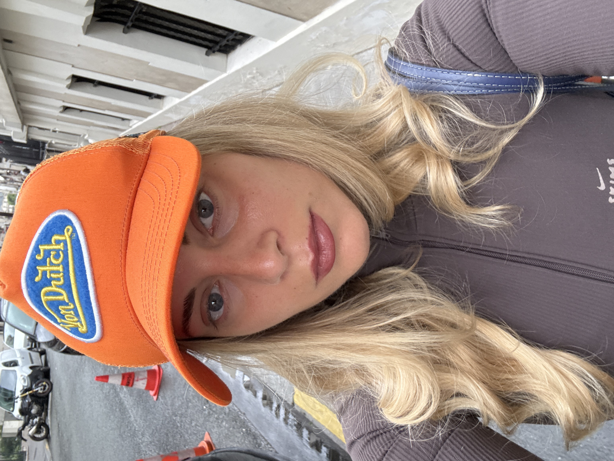 A woman poses in what appears to be Paris, France. The image has been taken at an angle, with the woman's face prominently in the foreground. She is wearing an orange Von Dutch cap. Her hair is long, wavy, and blonde. She is wearing a brown, ribbed zip-up top. In the background, there are Parisian-style buildings, and part of a road. Cones and a parked motorbike are visible on the street. The weather appears to be overcast or wet, judging from the sheen on the road.