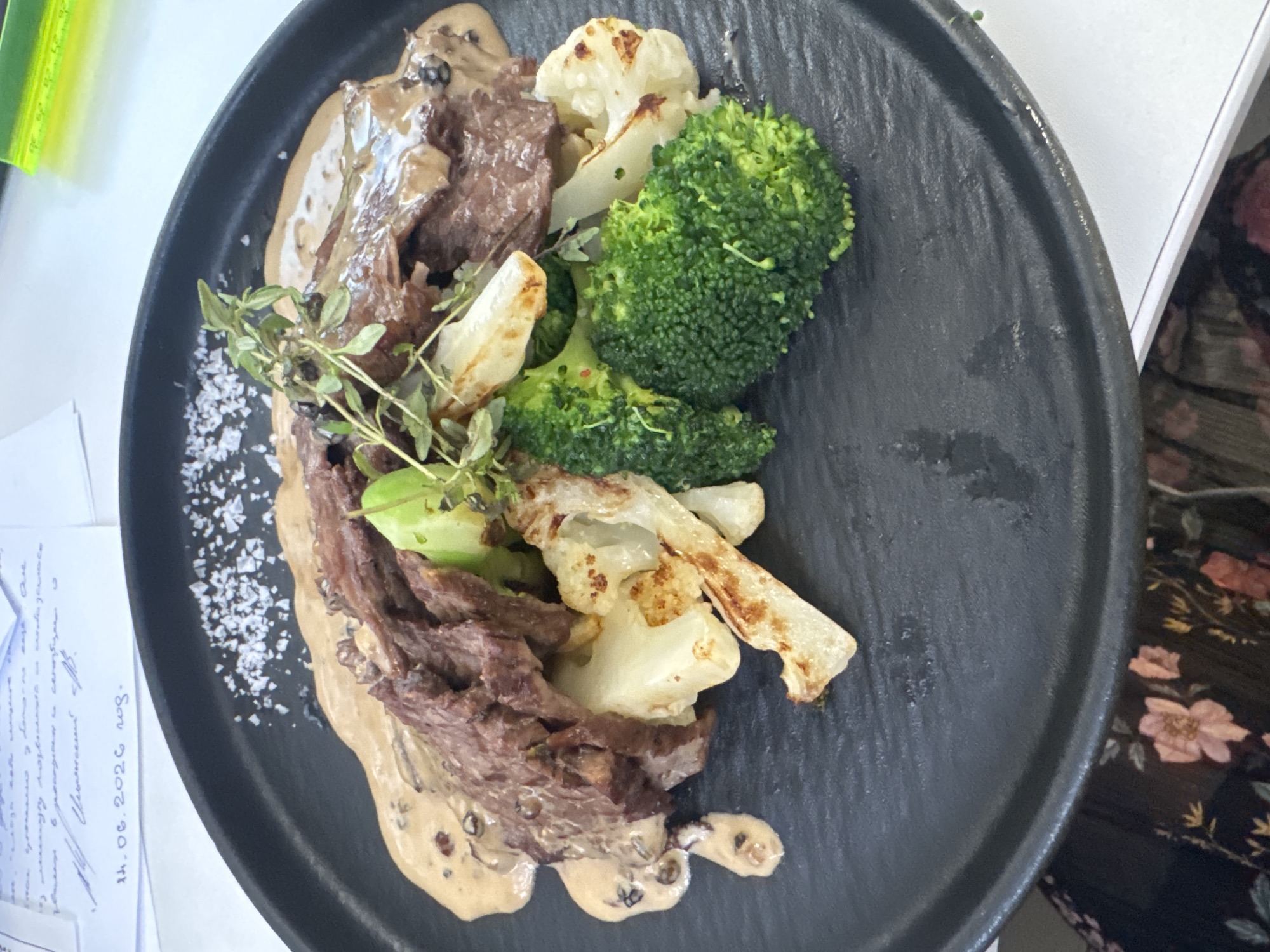 A close-up, eye-level shot captures a serving of steak with vegetables and sauce on a dark, textured plate. The steak, sliced into thin pieces, is draped over a mixture of roasted cauliflower and bright green broccoli florets. A creamy, speckled sauce is generously drizzled over the meat and vegetables, and a sprig of fresh thyme rests on top. Coarse salt crystals are sprinkled on the left side of the plate. In the background, a document with handwritten text and a date, "14.06.2026", is visible, along with a neon green ruler. The setting appears to be indoors, possibly a restaurant or a catered event. The image does not provide any specific details about the city of Moscow, Russia.