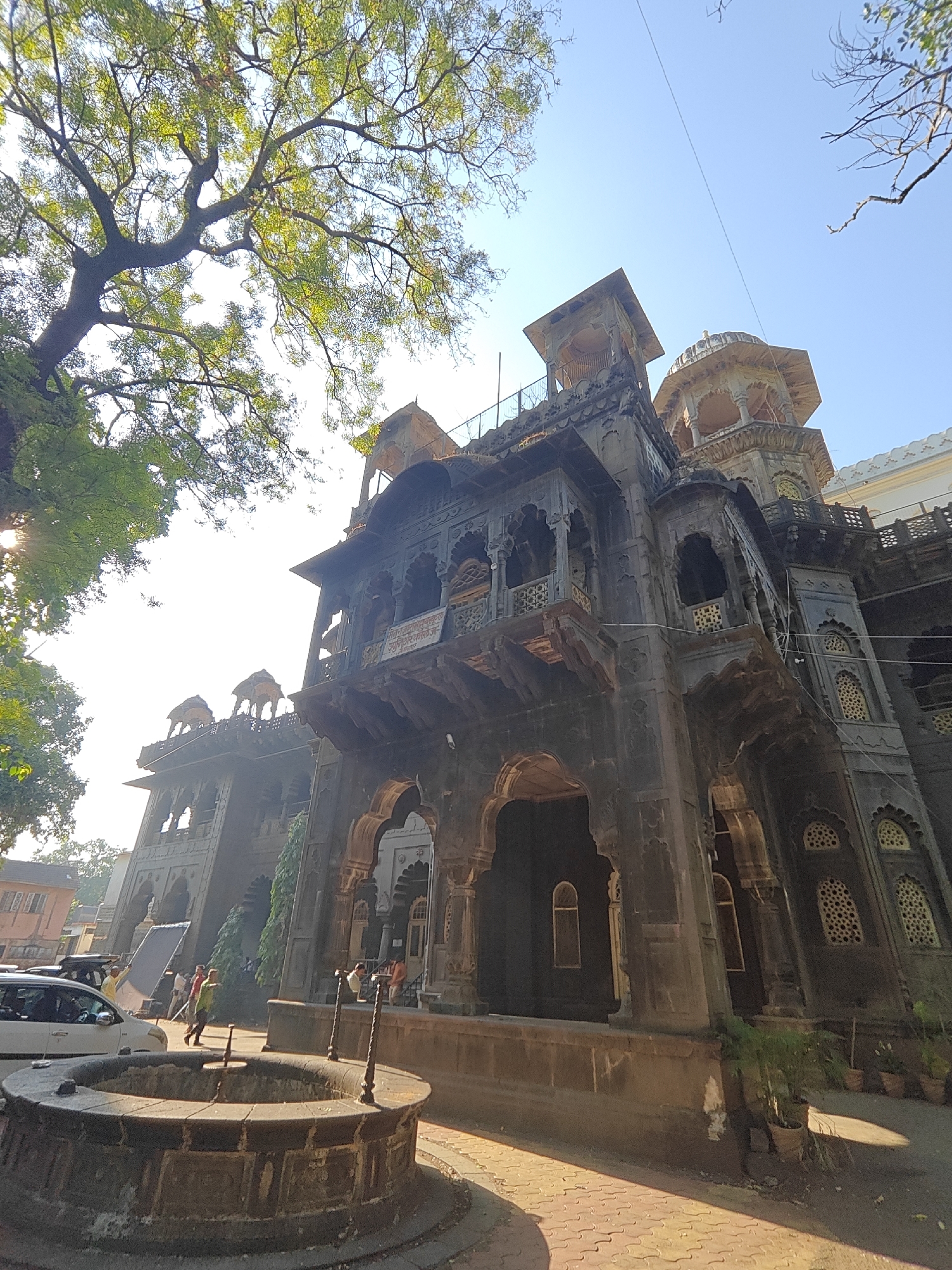 The image shows the New Palace in Kolhapur, India. It is a large, dark stone building with multiple stories, balconies, arches, and towers. The architecture features intricate carvings and designs. A large tree with green leaves is partially visible on the left side of the building, casting shadows. In front of the palace, there is a circular stone well. Several people are walking in the background near the entrance of the palace. A white car is parked to the left of the well. The sky is clear and blue. The ground is paved with bricks. There are small potted plants near the base of the building.