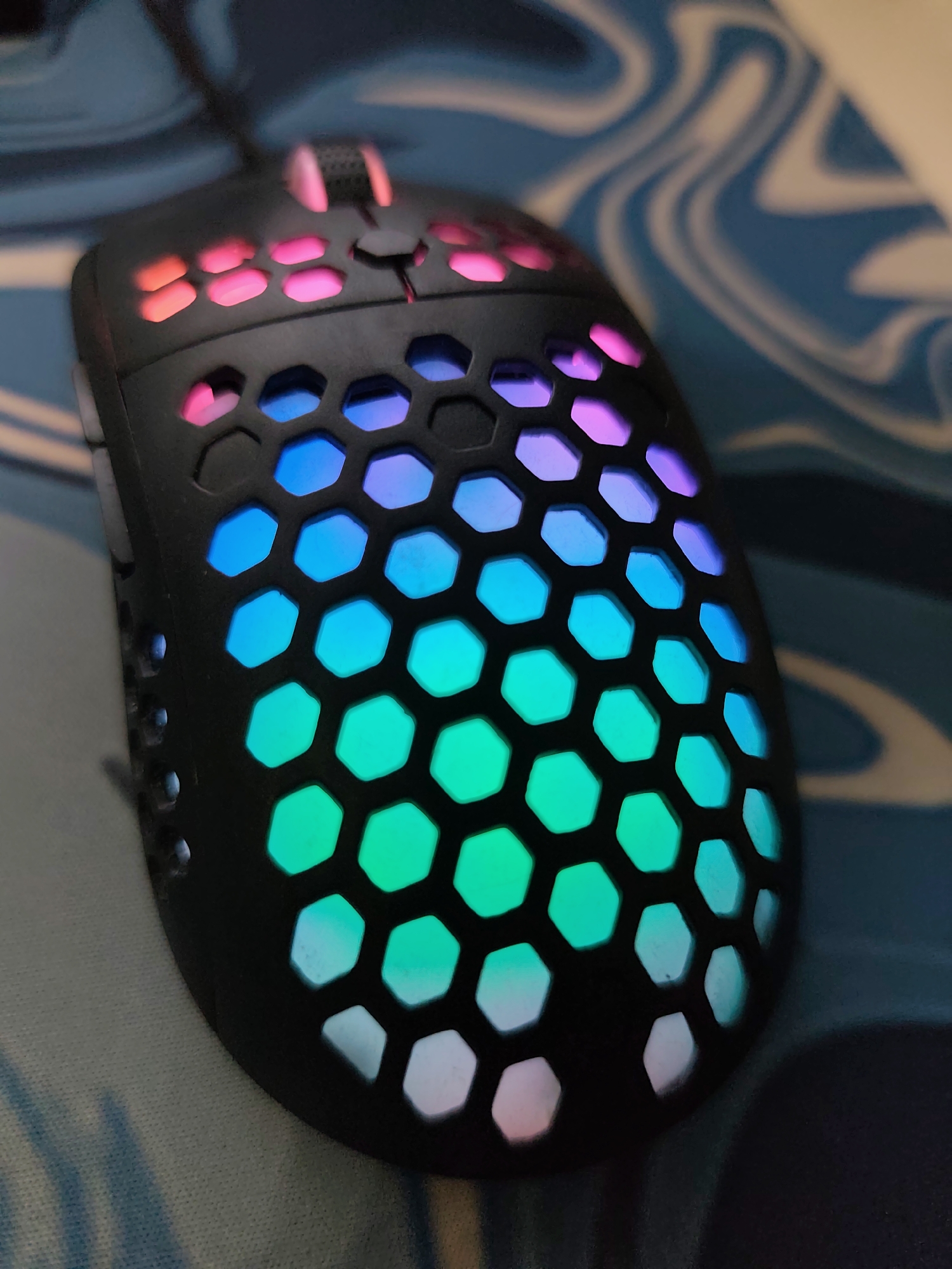 A black computer mouse featuring a hexagonal honeycomb-patterned shell is positioned on a blue and white patterned surface, likely a mousepad, in Pazardzhik, Bulgaria. The mouse is internally illuminated with RGB lighting, visible through the perforations. This lighting displays a color gradient from red and pink at the front, transitioning through blue and green, to white and light pink at the rear. A black scroll wheel with pink illumination is visible between the primary click buttons. A black cable extends from the front of the mouse. The background is out of focus, exhibiting abstract blue and white swirling patterns.