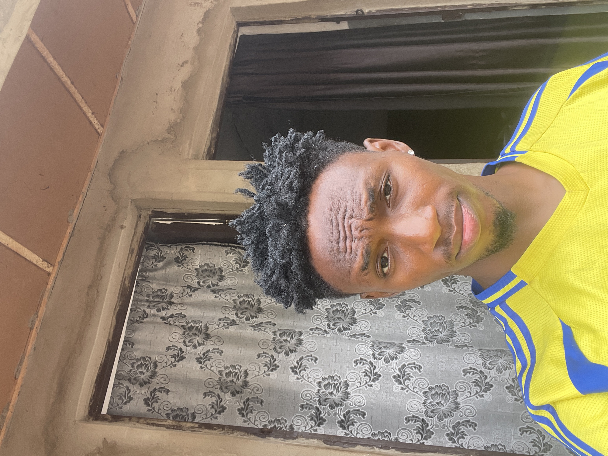 A medium-shot, eye-level photograph shows a Black man with dreadlocks, wearing a yellow and blue athletic shirt. He is positioned in front of a window covered with a patterned grey and black curtain. He has a slight beard, and his eyebrows are furrowed. A building exterior frames the top and left side of the image. A second window appears in the top center of the picture behind him. The photograph appears to have been taken at a skewed angle. The image may be located in Lokoja, Nigeria.