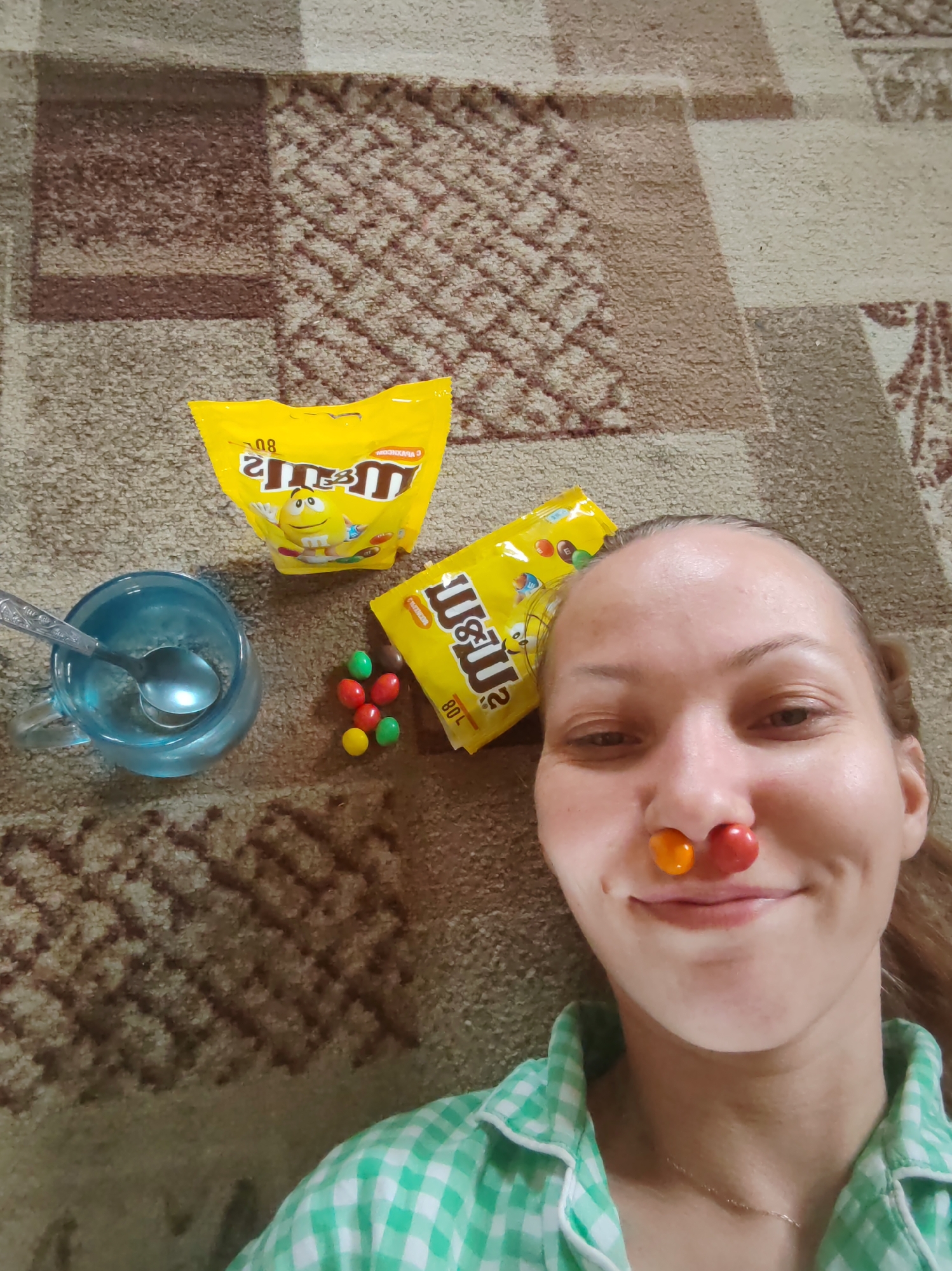 A woman lies on a brown patterned carpet with two bags of M&M candies, a clear mug with a spoon inside, and several loose M&M candies scattered nearby. The woman has placed an orange and a red M&M on her nose. She is wearing a green and white checkered shirt. The location is Чухлэм, Russia.