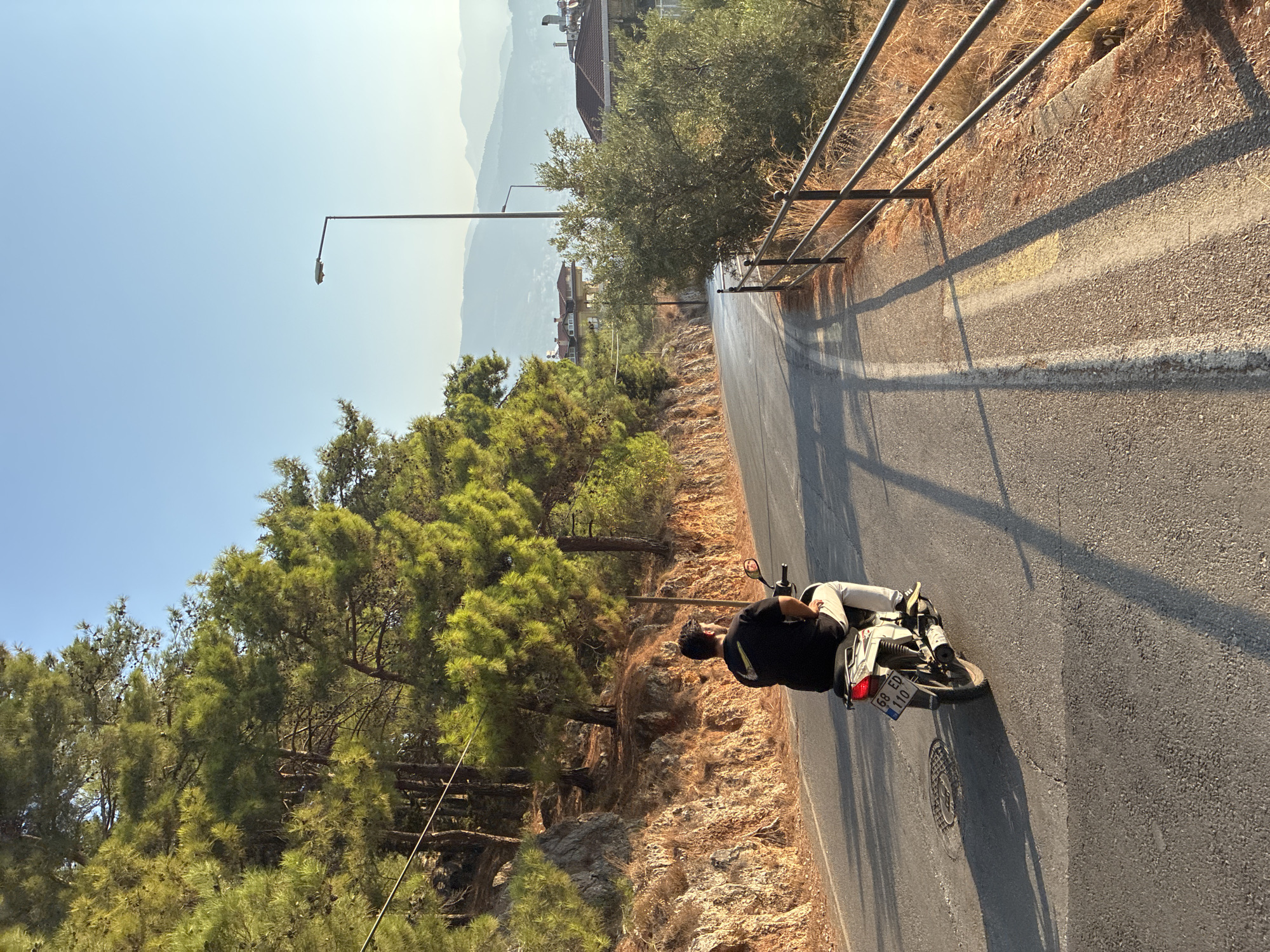 A man rides a scooter down a paved road lined with trees and dry vegetation. The road curves to the right, and in the distance, mountains are visible. A black metal fence runs along the right side of the road. The sun casts long shadows across the pavement. The sky is clear and blue. The license plate on the scooter reads "68 ED 110". The scene appears to be in a mountainous area on the outskirts of Alanya, Turkey.