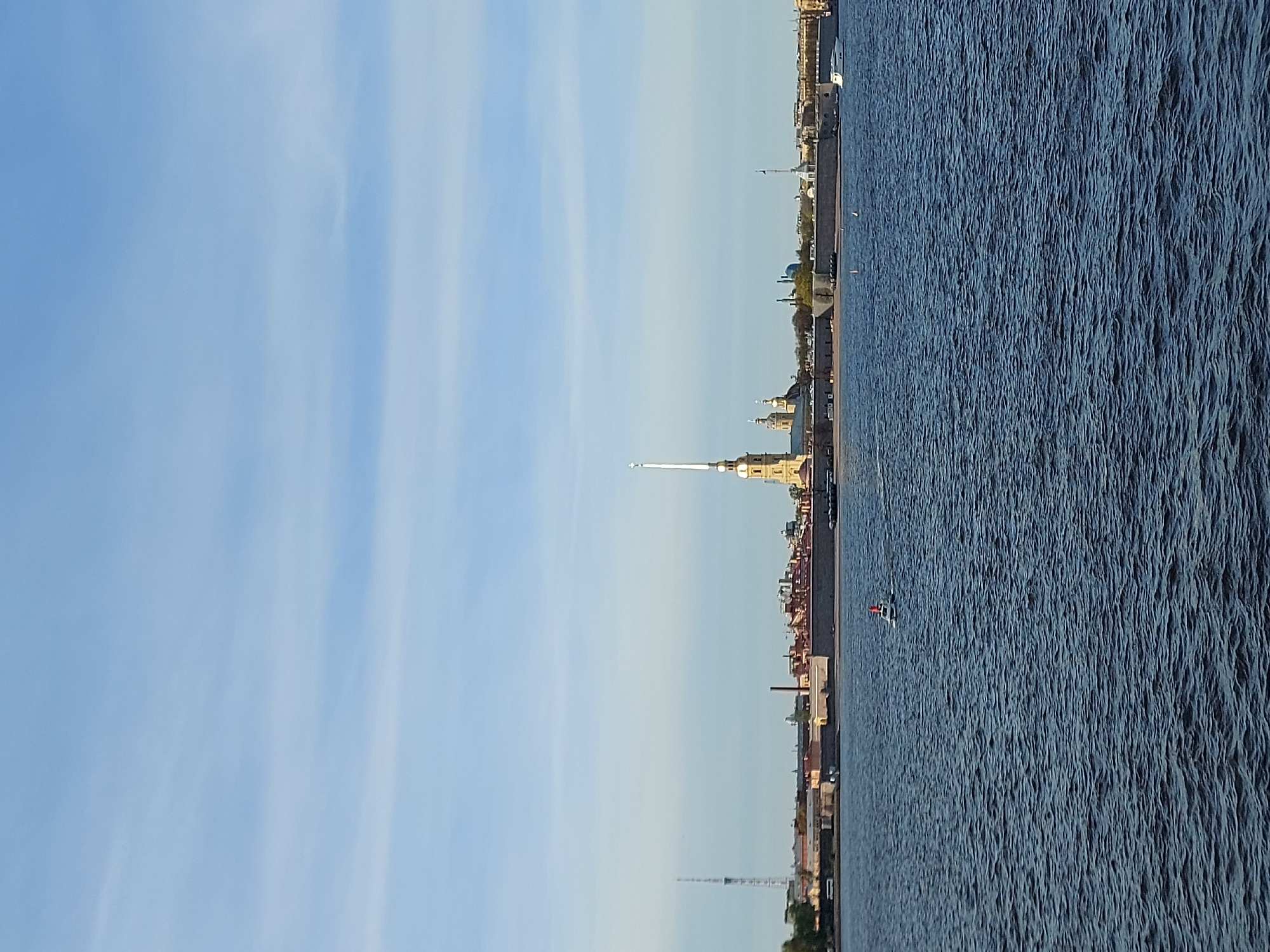 Here is a description of the image:
The image shows a cityscape of Saint Petersburg, Russia. The Peter and Paul Fortress is prominently featured, with its tall, slender spire reaching into the sky. A body of water occupies the right side of the frame, its surface textured by ripples and waves. A small boat is visible on the water. The sky, a pale blue, takes up a significant portion of the left side of the image. The image is oriented vertically.
