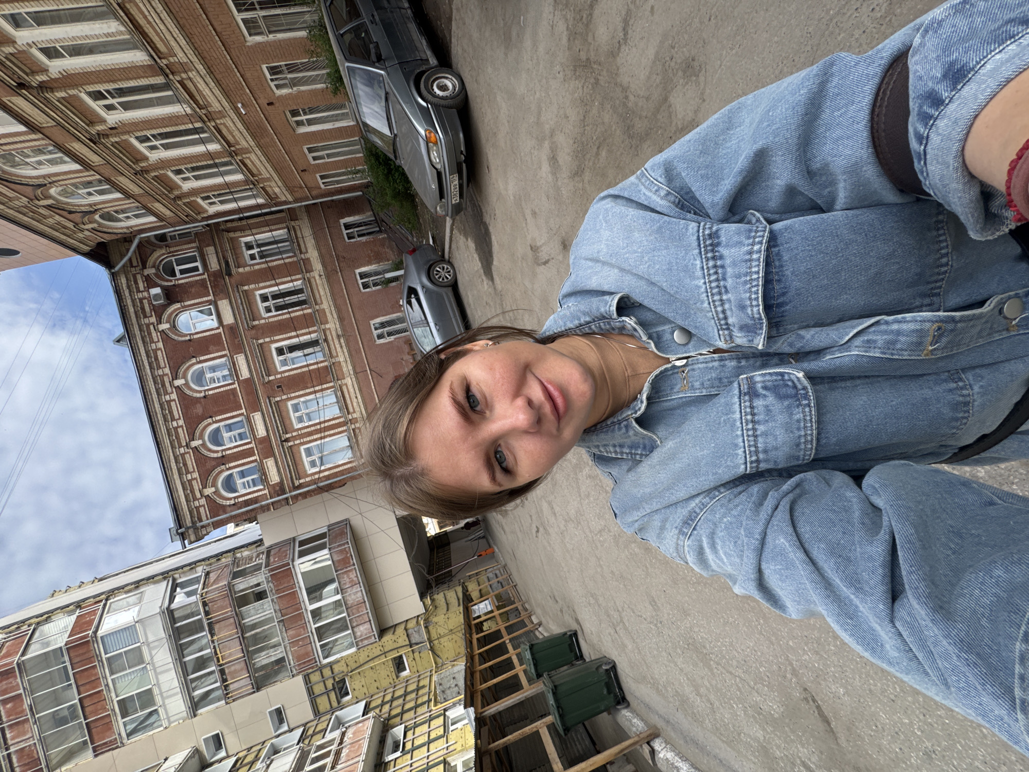 A selfie captured in Perm, Russia, shows a blonde-haired woman wearing a blue denim jacket standing outside. The backdrop features two adjacent buildings. One is a brick building with multiple floors, distinguished by a repetitive pattern of windows with arched tops on certain floors. Adjacent to it is a more modern building under construction, showing exposed insulation panels and scaffolding. Two parked gray cars are visible, one closer to the older brick building. The scene is set on a paved road or walkway.