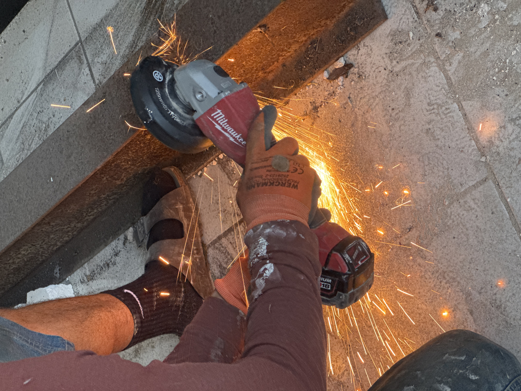 An individual, partially visible from the waist down, is using a red and gray Milwaukee angle grinder to cut into a metal beam. The individual is wearing a burgundy long-sleeved shirt, blue denim shorts, dark socks with a white Nike logo, and orange work gloves. Sparks are flying from the grinding action, illuminating the concrete floor and the metal beam. The environment appears to be outdoors, possibly in a construction or workshop area, with tiled flooring visible in the background. The image captures a moment of intense activity, highlighting the danger and precision involved in metalworking.