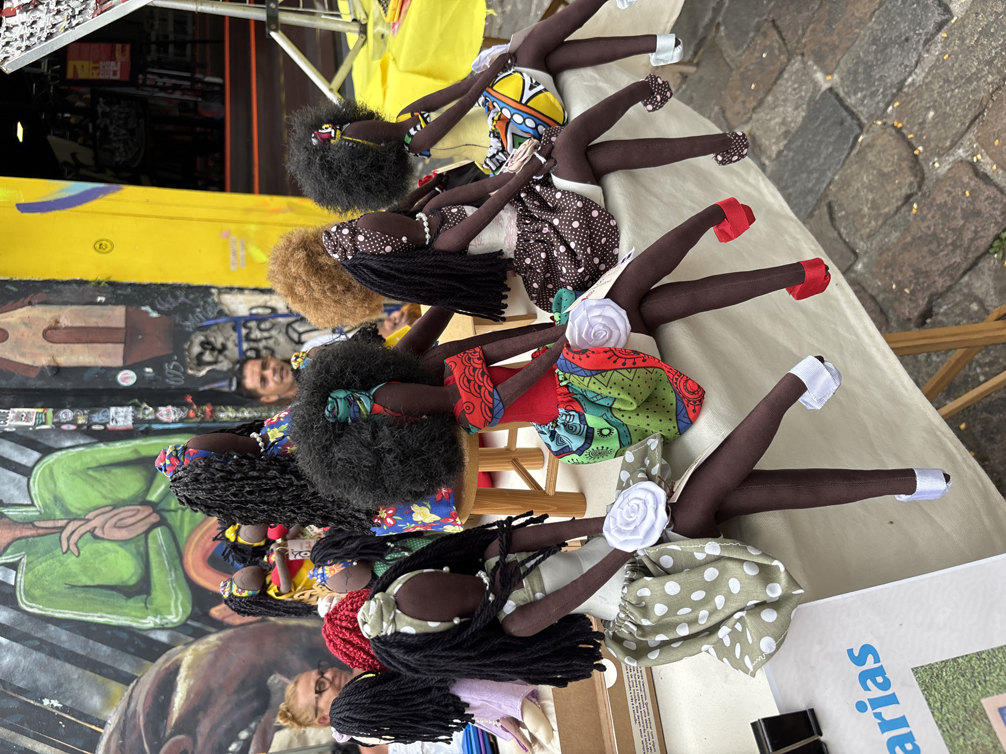 A street scene in São Paulo, Brazil, features a display of handmade dolls. The dolls are lined up on a table with a light-colored tablecloth. Each doll has a dark complexion and is dressed in vibrant, patterned clothing. They have distinct hairstyles, including afro puffs and long braids. A small wooden stool is positioned amongst the dolls. The background includes a colorful graffiti-covered wall. To the right, part of a document with the word "arias" is visible. The setting suggests a market or craft fair within the urban environment.