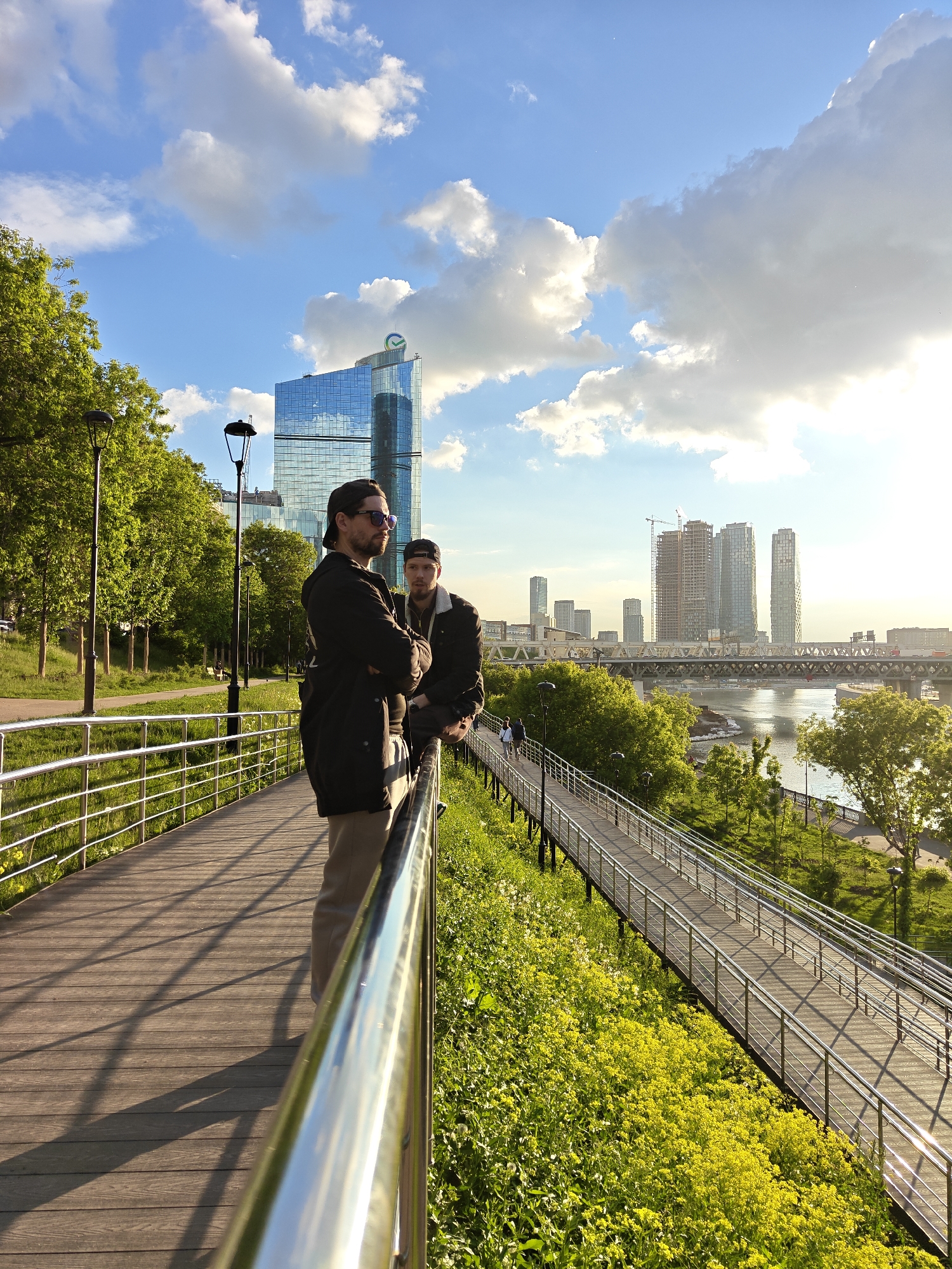 Two men are standing on a wooden boardwalk with metal railings. The man in the foreground is wearing sunglasses, a dark jacket, and light-colored pants. He has his arms crossed. The second man, slightly behind, wears a cap and dark jacket.
The boardwalk runs parallel to a grassy slope covered in yellow flowers. Further down, another boardwalk descends toward the river. In the background, a cityscape includes modern skyscrapers and a bridge over the river. The scene is bathed in sunlight under a blue sky with scattered clouds.
The location is Moscow, Russia, with Moscow International Business Center (Moscow City) in the background.