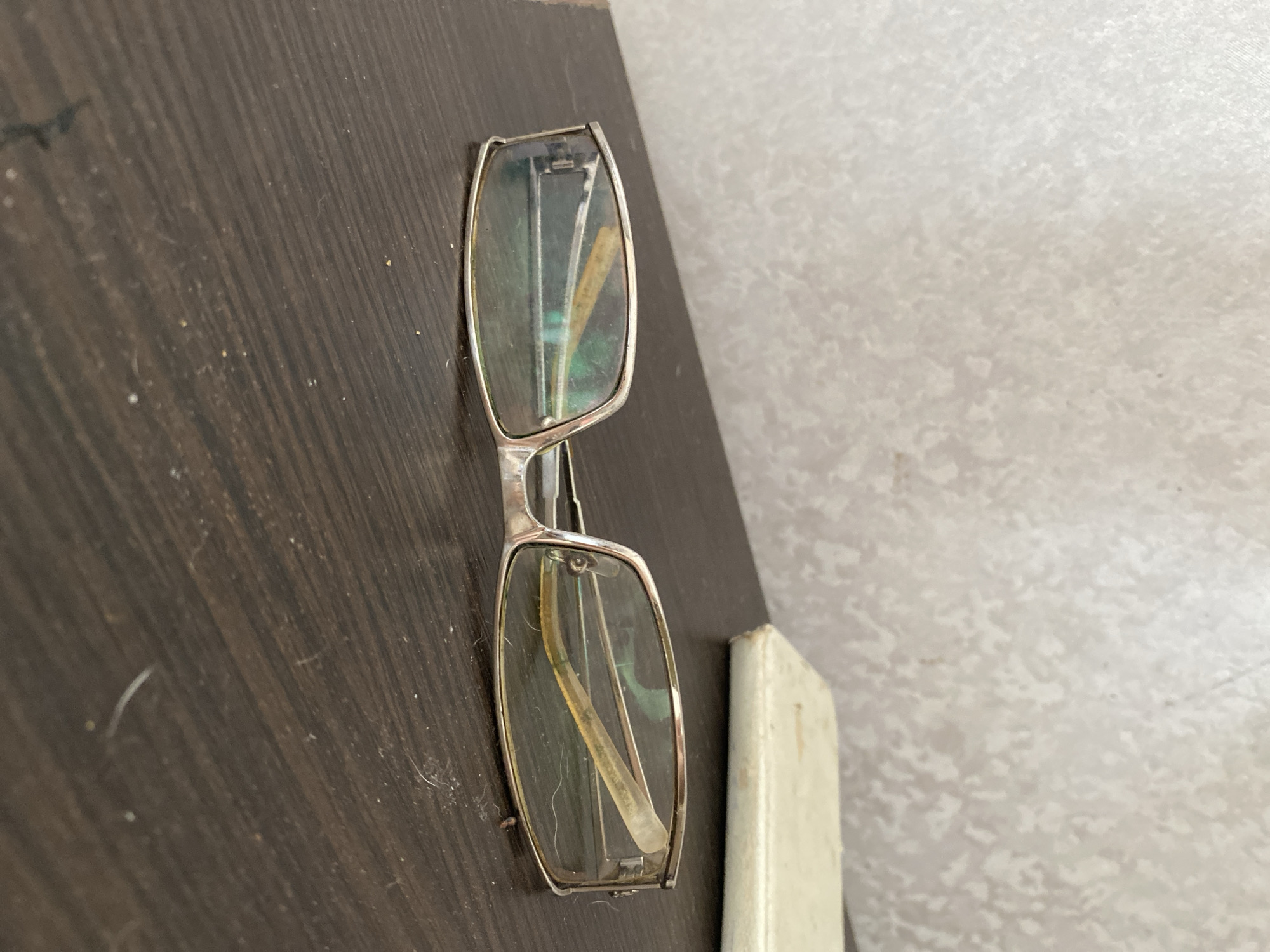 An image shows a pair of eyeglasses lying on a dark, wooden surface. The eyeglasses have thin, silver-colored frames and clear lenses that appear to have some smudges and reflections. The background is a textured, off-white wall. To the right of the eyeglasses, a pale, rectangular object, possibly a case, is partially visible. The overall scene appears to be indoors. The provided information that this image is located in the city of Chornomorsk, Ukraine, is not directly evident from the visual content of the image itself.