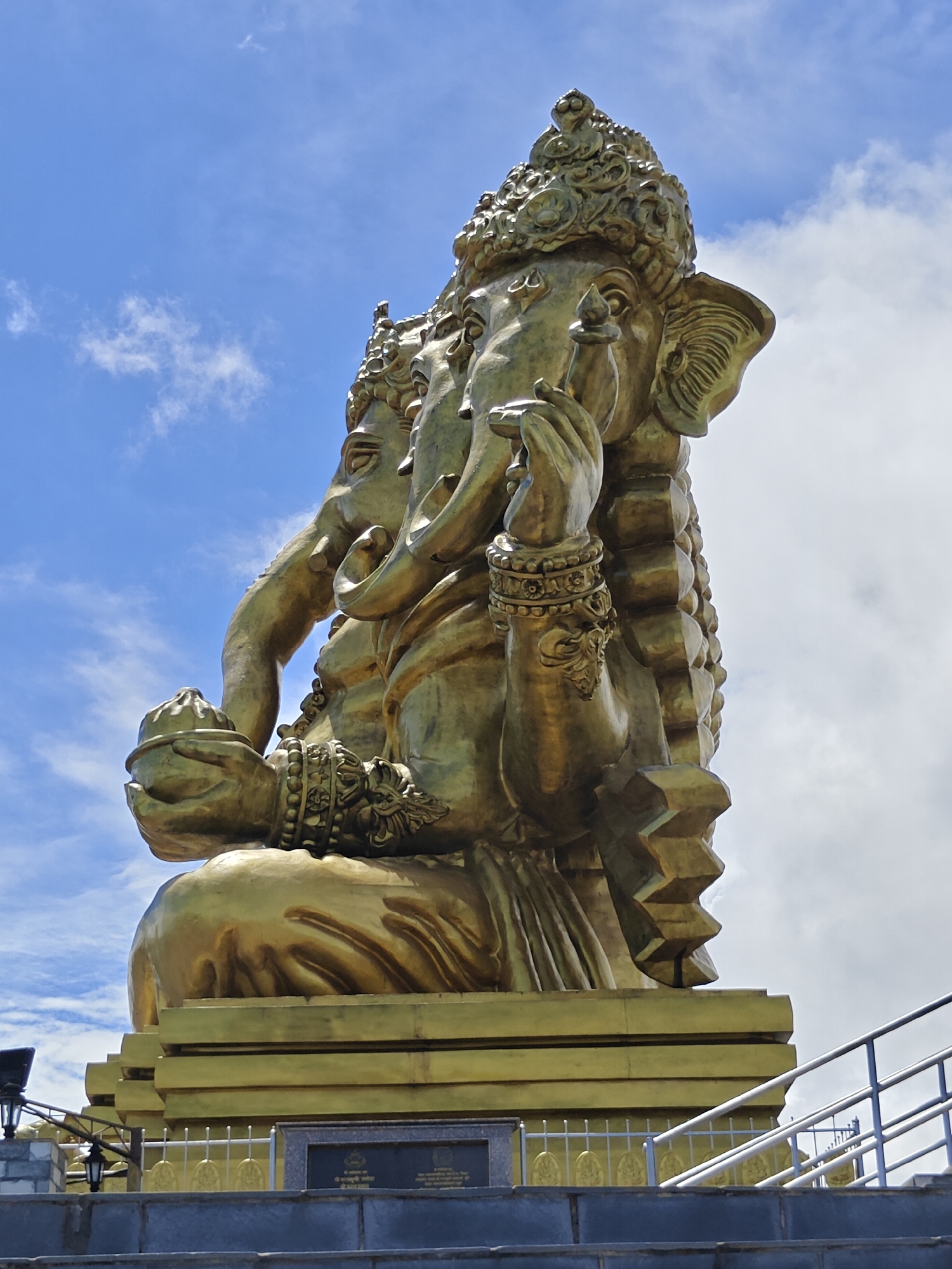 The image shows a low-angle, medium shot of the gigantic golden statue of Lord Ganesha, a prominent Hindu deity, situated at the top of a hill in Pokhara, Nepal. The statue is depicted in a seated position, with multiple faces and arms, and is richly adorned with intricate jewelry and headdresses. The statue is made of brass, reflecting the sunlight and creating a lustrous golden hue.
The statue is perched on a multi-tiered, golden pedestal. In the foreground, a set of stone steps leads up towards the statue, with metal railings on either side. A plaque with inscriptions is visible on the pedestal. In the background, a bright blue sky with scattered white clouds forms the backdrop. The overall impression is one of grandeur and spiritual significance, likely a popular tourist and pilgrimage site.