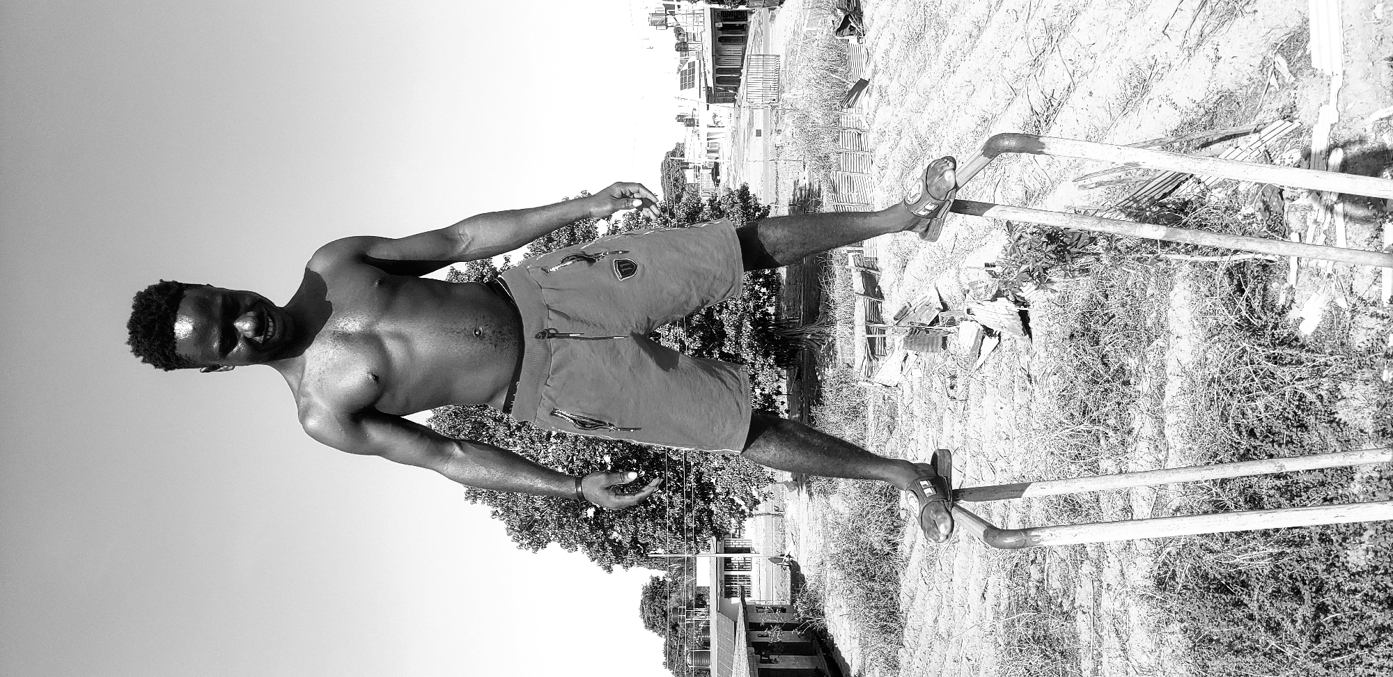 A black and white image shows a shirtless dark-skinned man standing on two parallel metal bars. He is wearing shorts and sandals. The location appears to be outdoors, possibly in Narayi, Nigeria, given the prompt. The background consists of sparse vegetation and indistinct buildings in the distance. There are no other people present. The man is the primary subject, and his interaction with the bars suggests a form of exercise or play.