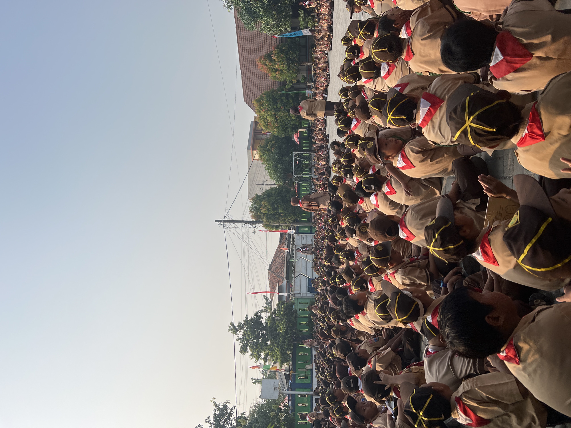 An outdoor assembly of young people, likely students in scout uniforms, is depicted. The individuals are seated on the ground in rows, facing towards the center of the gathering where two adult figures stand. The uniforms consist of light brown shirts and brown hats adorned with yellow criss-cross patterns. Red neckerchiefs are visible on many of the younger participants. The environment appears to be a schoolyard or open field, with buildings and trees in the background. Indonesian flags, red and white, are displayed on poles, suggesting a national event or ceremony. The lighting indicates it is daytime, with the sun casting a warm glow on the scene. The participants are predominantly young males and females, indicating a mixed-gender event. The gathering seems orderly, with participants attentive to the individuals speaking.