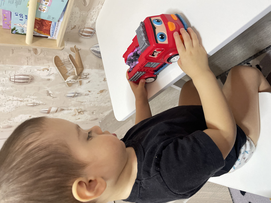 A young child is seated at a white table, positioned in what is identified as Izhevsk, Russia. The child, with light brown hair and fair skin, wears a black short-sleeved t-shirt and a patterned diaper. Their attention is directed downward and to the right, focusing on a red toy fire truck situated on the table directly in front of them. Both of the child's hands are on the toy; one hand rests on the front of the truck, near its cartoonish face, and the other hand is placed on the side of the vehicle.
The toy fire truck is predominantly red with blue and orange accents, featuring large blue eyes, a smiling mouth, and an image of a firefighter in a side window. A segment of purple and grey material is visible in the rear compartment of the truck.
The wall behind the child and table is covered with a light-colored wallpaper featuring an illustrated world map with various modes of transportation such as hot air balloons and biplanes. To the right, a light wooden wall-mounted shelf holds several books, one of which displays Cyrillic text. A segment of a patterned rug or carpet with grey, white, and black elements is visible on the floor beneath the table.