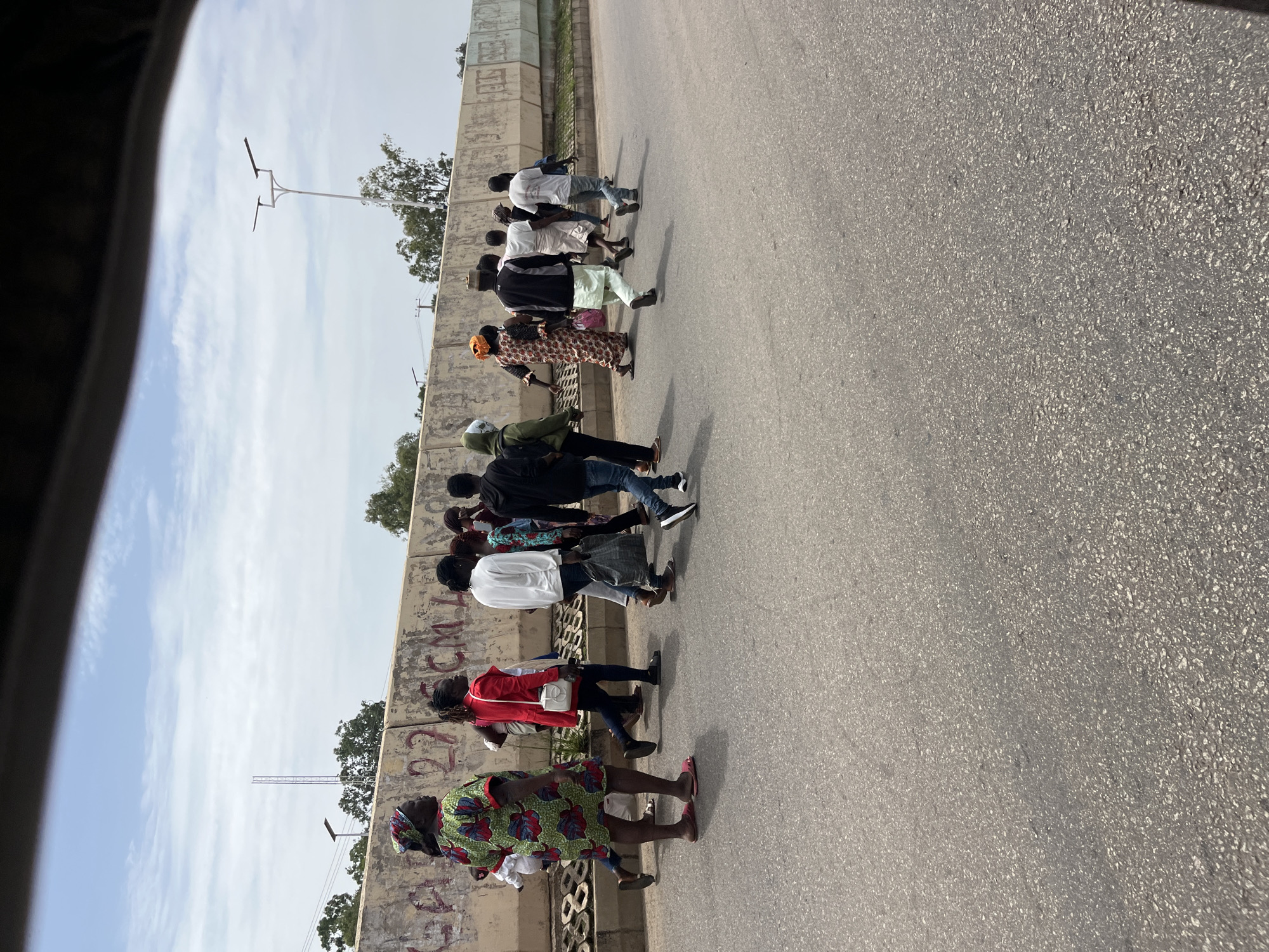A group of people are walking along a street in Jos, Nigeria. The group consists of both men and women, dressed in casual clothing. Some individuals are wearing headscarves, suggesting a cultural context. The ground is paved with asphalt, and there is a concrete wall with graffiti on the left side of the image. Trees and utility poles are visible in the background, indicating an urban or suburban setting. The sky is partly cloudy, suggesting daytime with potentially warm weather. The people appear to be walking in the same direction, possibly heading to a destination or simply passing by. There is no specific event or activity clearly depicted, other than pedestrian movement. The lighting and shadows suggest it is late morning or early afternoon. The graffiti on the wall is in an illegible script, making it impossible to discern any specific text or message. The overall scene is ordinary, depicting everyday life in an African city.