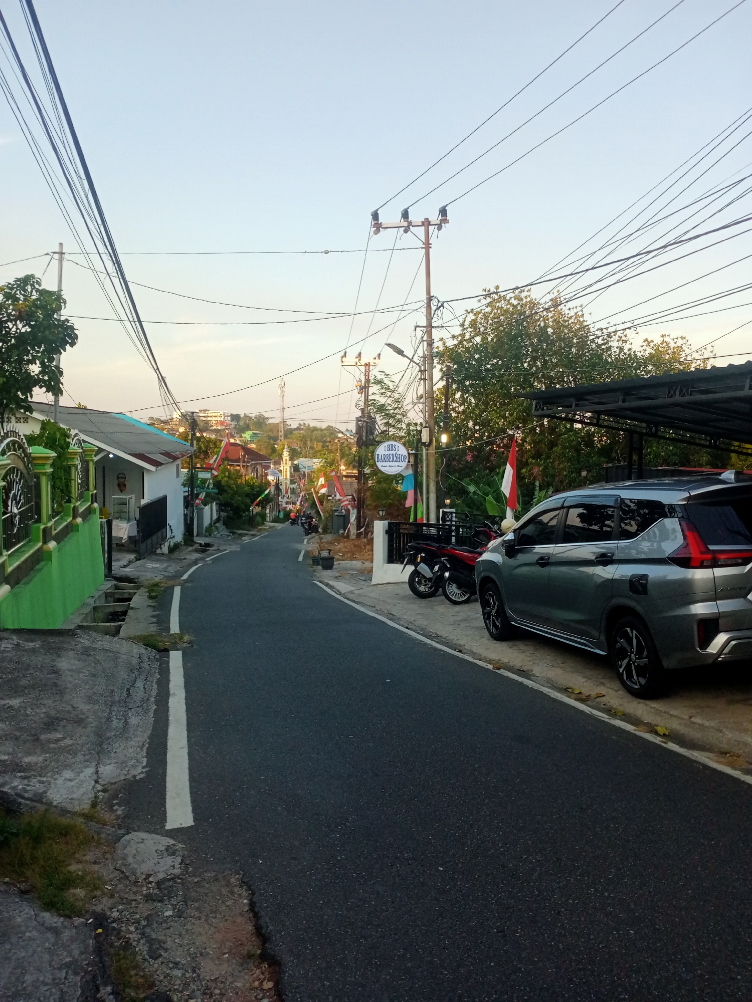 The image depicts a street scene in Balikpapan, Indonesia. A gray SUV is parked on the right side of the road, partially under a covered structure. To the left of the SUV, two motorcycles are parked. A sign for "BBS BARBERSHOP" is visible on a pole. Indonesian flags are displayed along the street. The road is asphalt and curves uphill. Houses and trees line the street. Overhead, a tangle of power lines crisscrosses the sky. The lighting suggests it is either early morning or late afternoon.