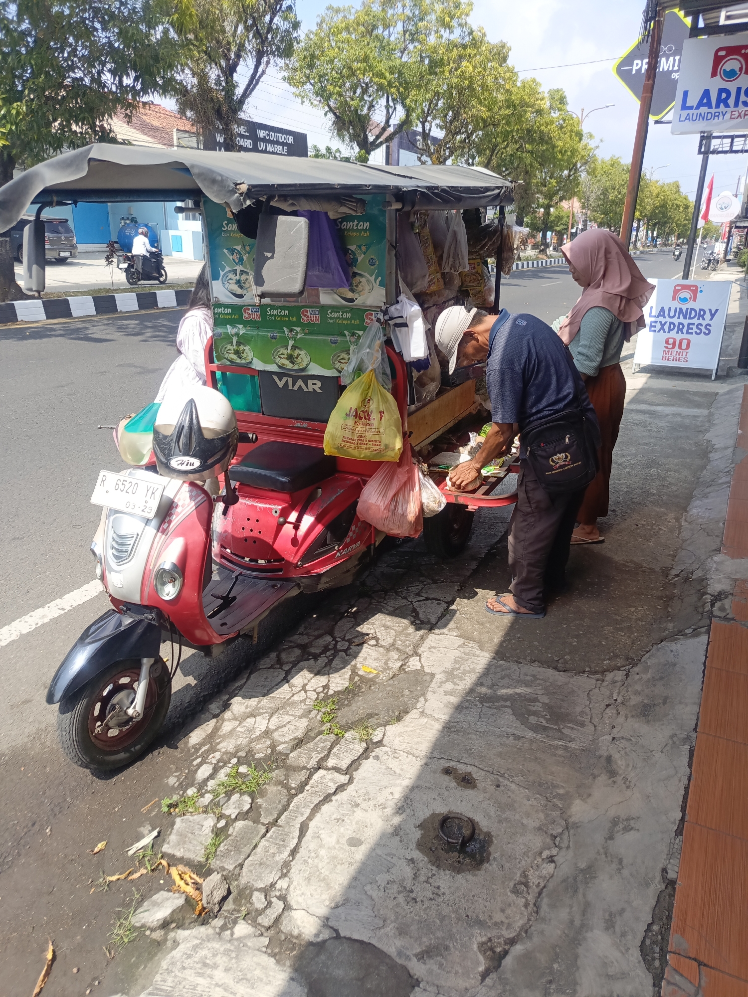 A red and white motorized tricycle food cart is parked on the side of a paved road in Desa Gunungsimping, Indonesia. A man wearing a blue shirt, grey pants, a white cap, and flip-flops is standing behind the cart, preparing food. A woman wearing a light brown hijab and a light green shirt is standing to the right of the man, looking at the cart. The food cart has a dark canopy and is decorated with green banners and images of food. There are plastic bags containing food items hanging from the cart. A motorcycle with a license plate is parked on the left side of the frame, and a laundry service sign is visible on the right. The ground is a mixture of cracked concrete and asphalt with patches of grass. Trees line the opposite side of the road.