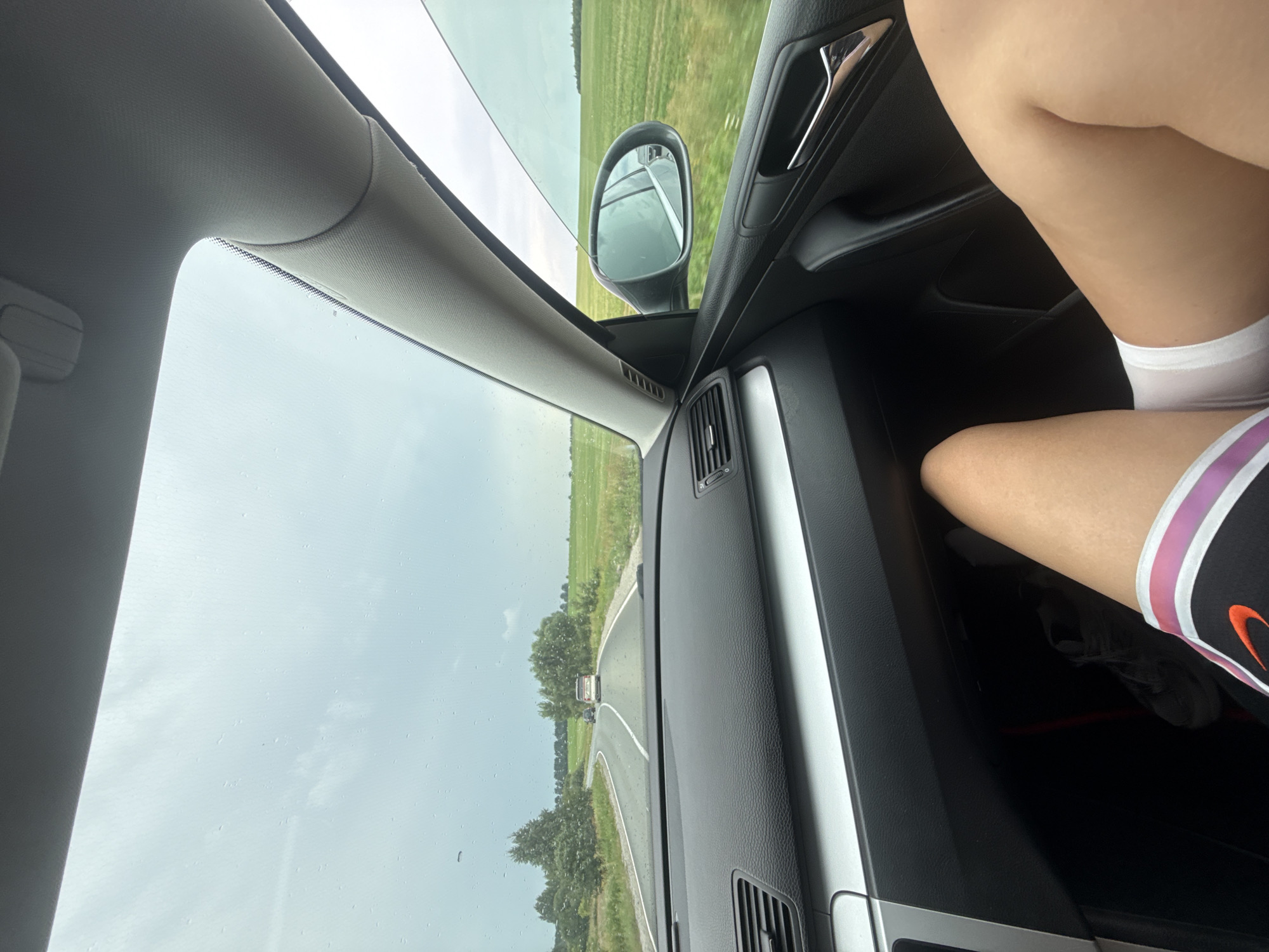 The image shows a view from inside a car driving on a road in Perevoznoe, Russia. The sky is overcast with some clouds, and there are raindrops visible on the windshield. The road is asphalt with a white line marking the lanes. On either side of the road are green fields and trees. In the distance, there are other cars on the road. Inside the car, the dashboard and passenger seat are visible. A person's legs are also visible, wearing white shorts and athletic shoes. The overall scene depicts a drive through the countryside on a cloudy day.