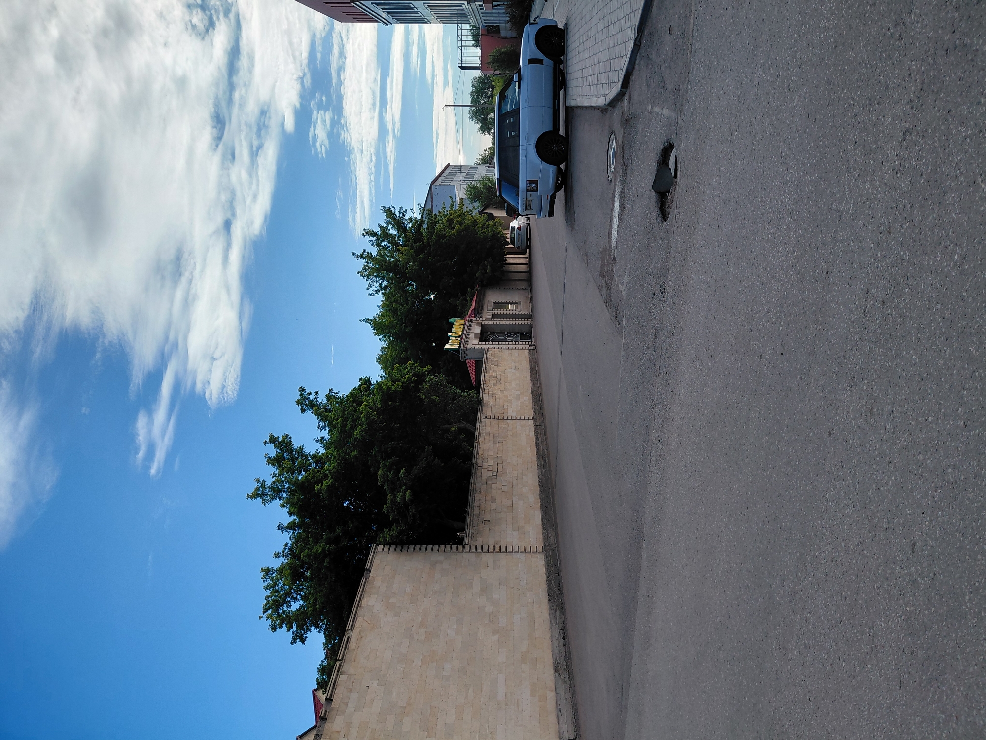 A light blue Range Rover is parked on the side of a paved road in Karaganda, Kazakhstan. The road has several potholes. To the left of the road is a long, low, beige brick wall topped with a darker colored coping. Behind the wall, trees are visible. A sign that reads "Kids Garden" is mounted above an arched entrance in the wall. Further down the street, a multi-story building is visible. To the right of the road, a modern building with a red facade is present. The sky is blue with scattered white clouds.