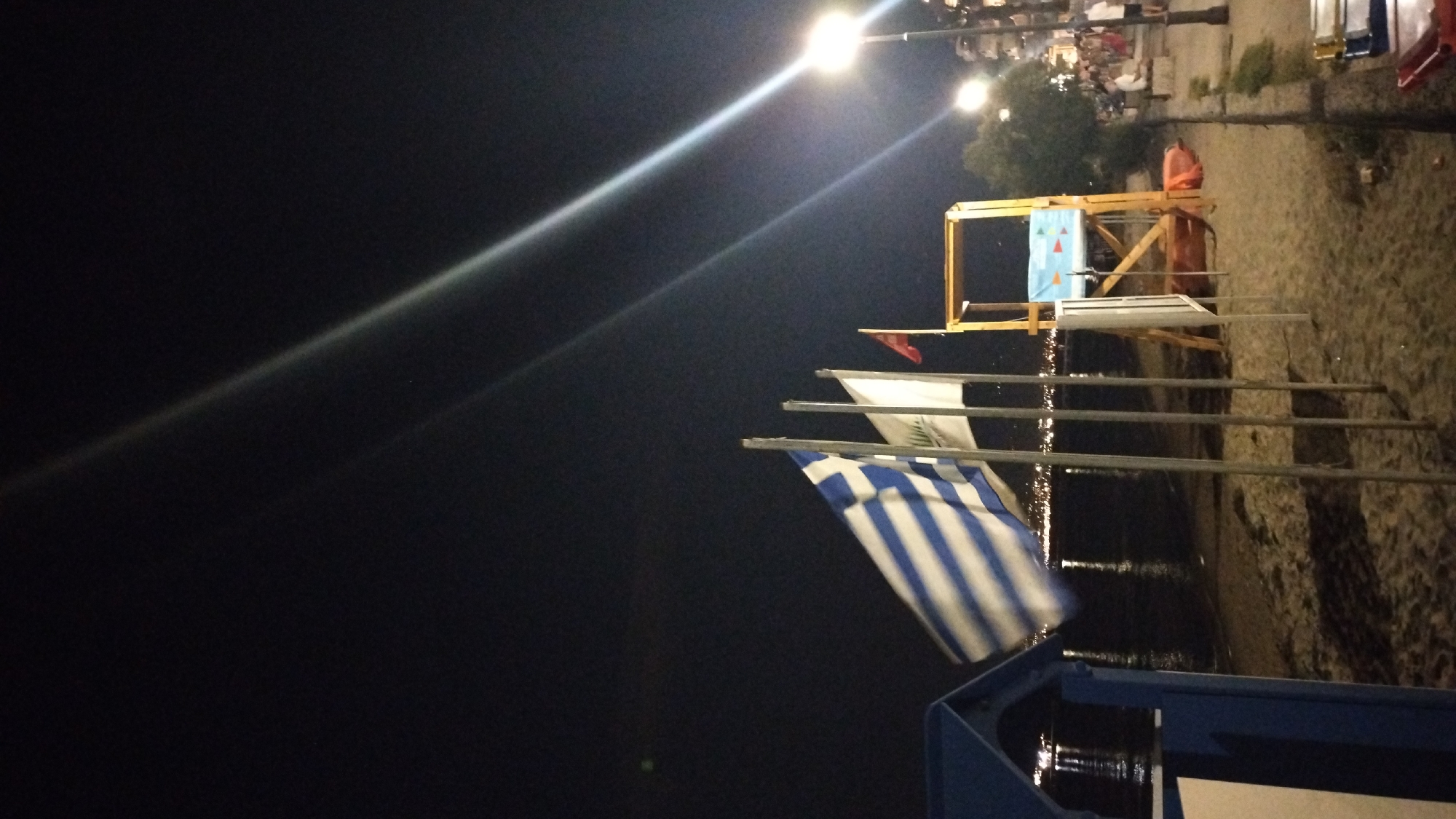 At night, on a sandy beach in Perea, Greece, several flags are displayed on poles. The foremost flag is the Greek flag, identifiable by its blue and white stripes and a white cross on a blue canton. To its right, two other flags are visible: a white flag, possibly bearing a logo or emblem, and a small red flag. In the background, a lifeguard tower made of wood stands on the sand, with a blue and white banner attached to its front. The sea is to the left of the beach, with lights reflecting on its surface, suggesting a town or city nearby. The sky is dark, with a bright light beam extending from the upper right corner of the image, likely from a street lamp. Several people are scattered on the beach and on a paved walkway to the right, indicating a public area. A blue structure, possibly a beach hut or changing room, is partially visible on the far left.